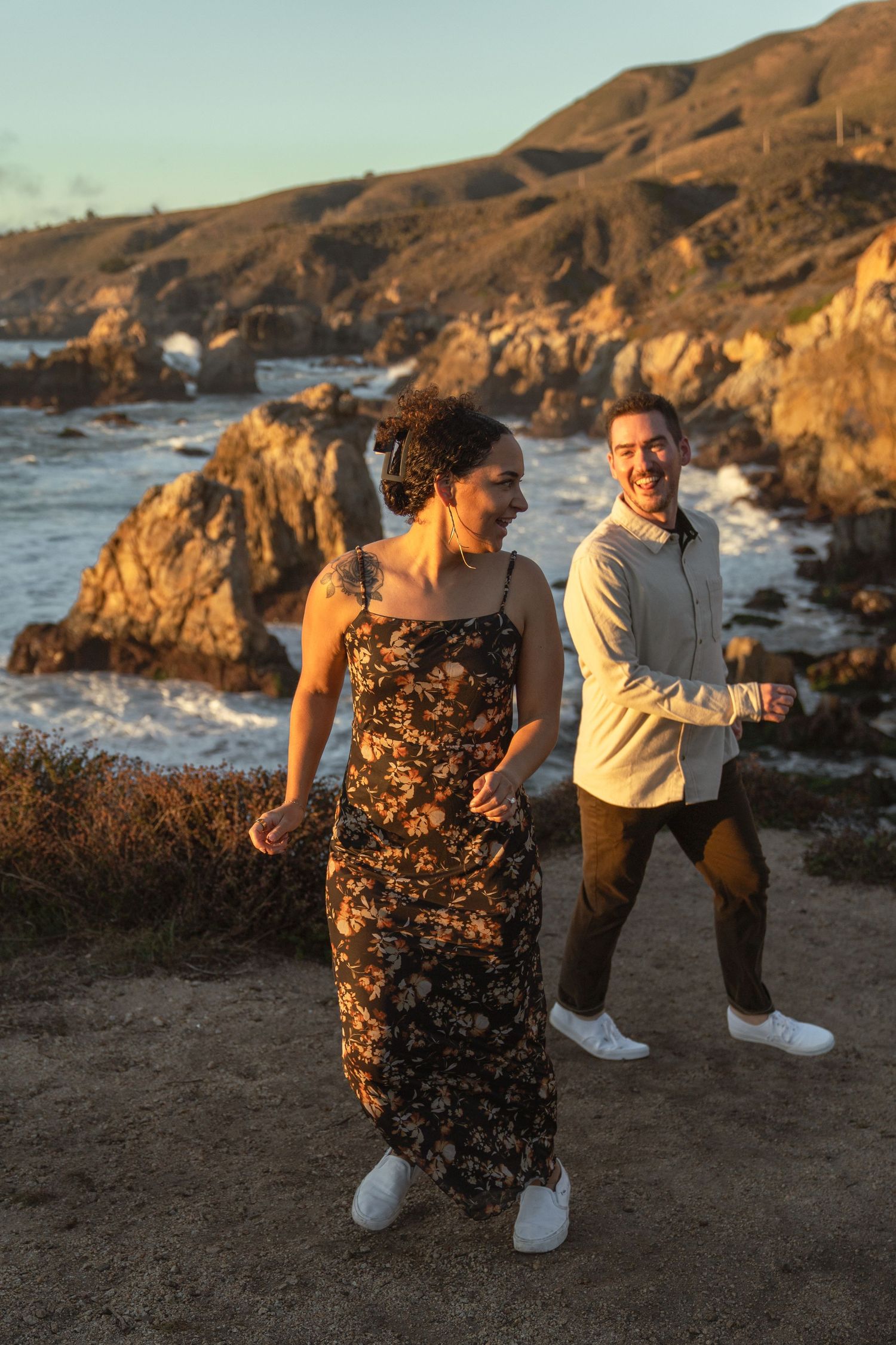 A couple shares a playful moment at sunset along a rocky coastal cliff with ocean views in the background.