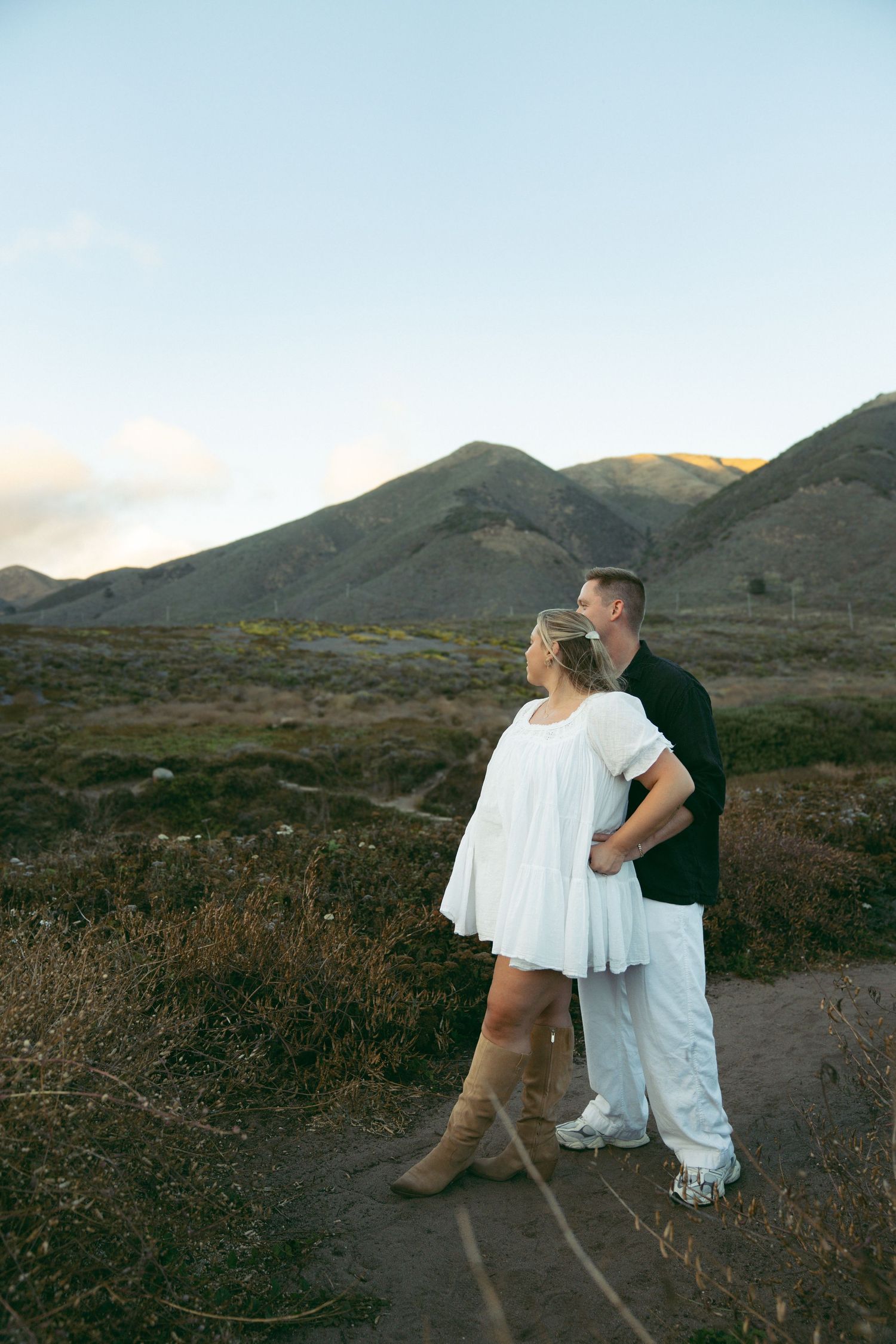 Couple embracing on a mountain trail during sunset with scenic coastal views in the background.
