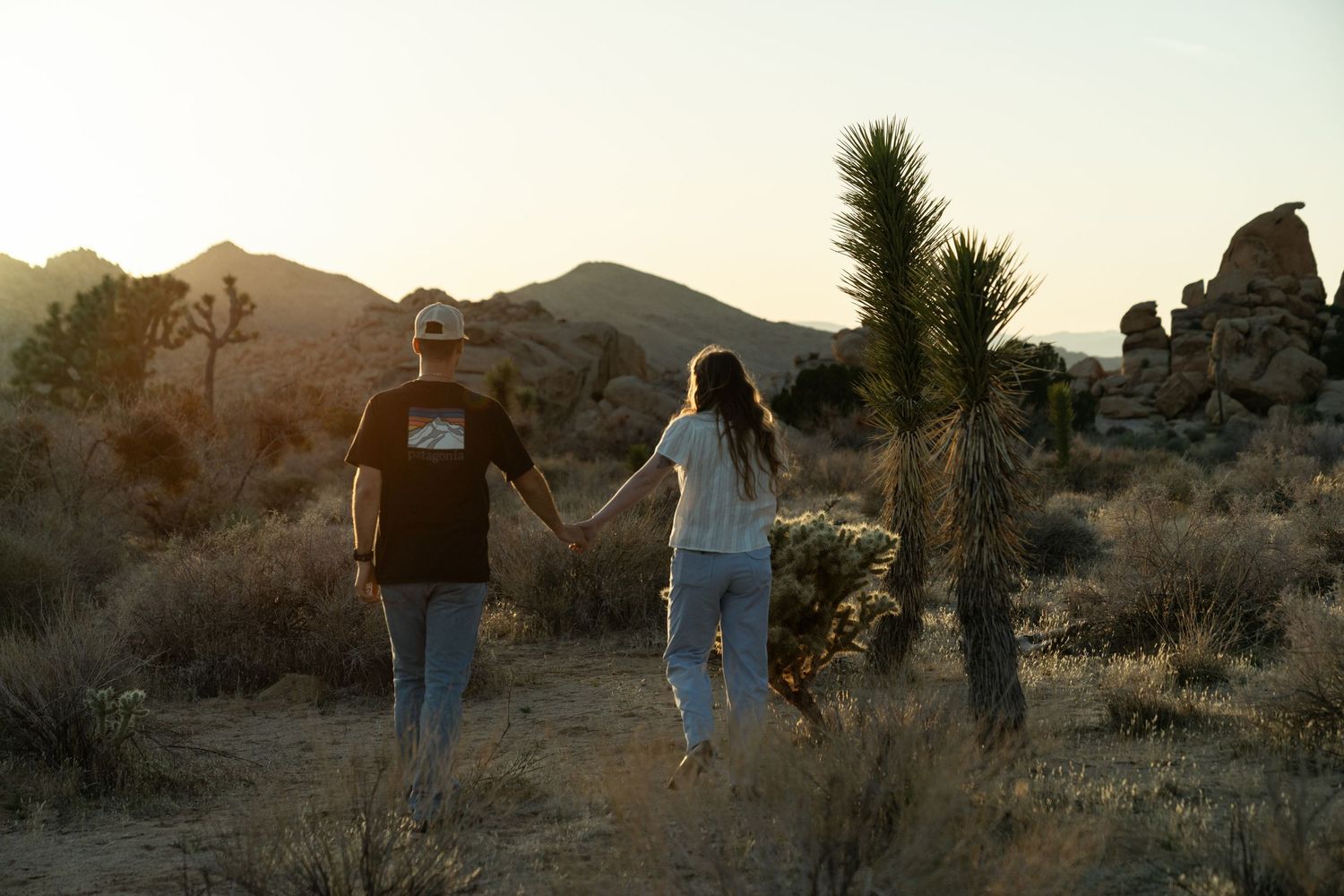 A couple explores a desert landscape against the backdrop of rocky terrain at sunset.