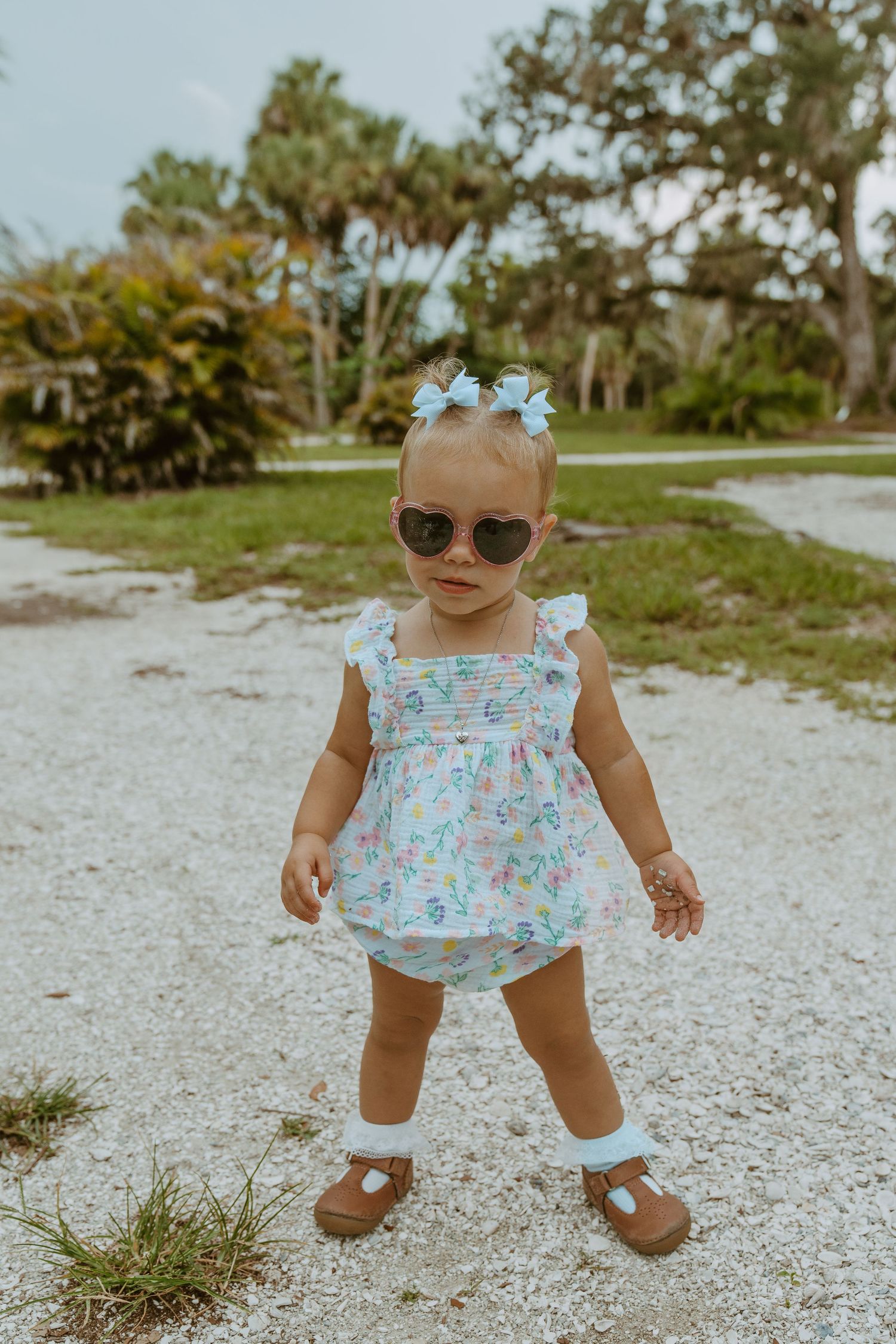 Toddler in floral dress and sunglasses standing confidently on gravel path.