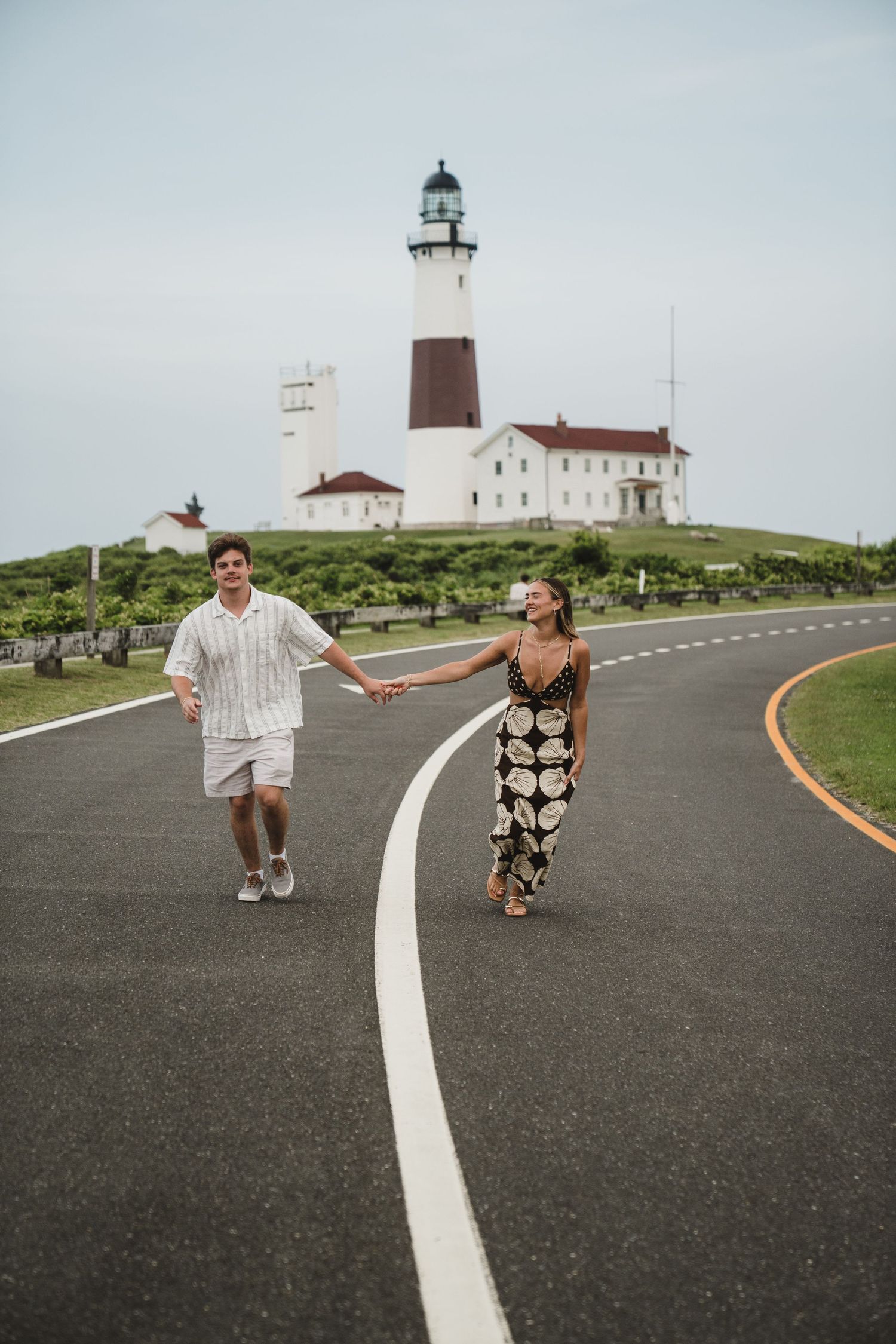 Several connected photos of people walking on a path towards a lighthouse on a cloudy day.