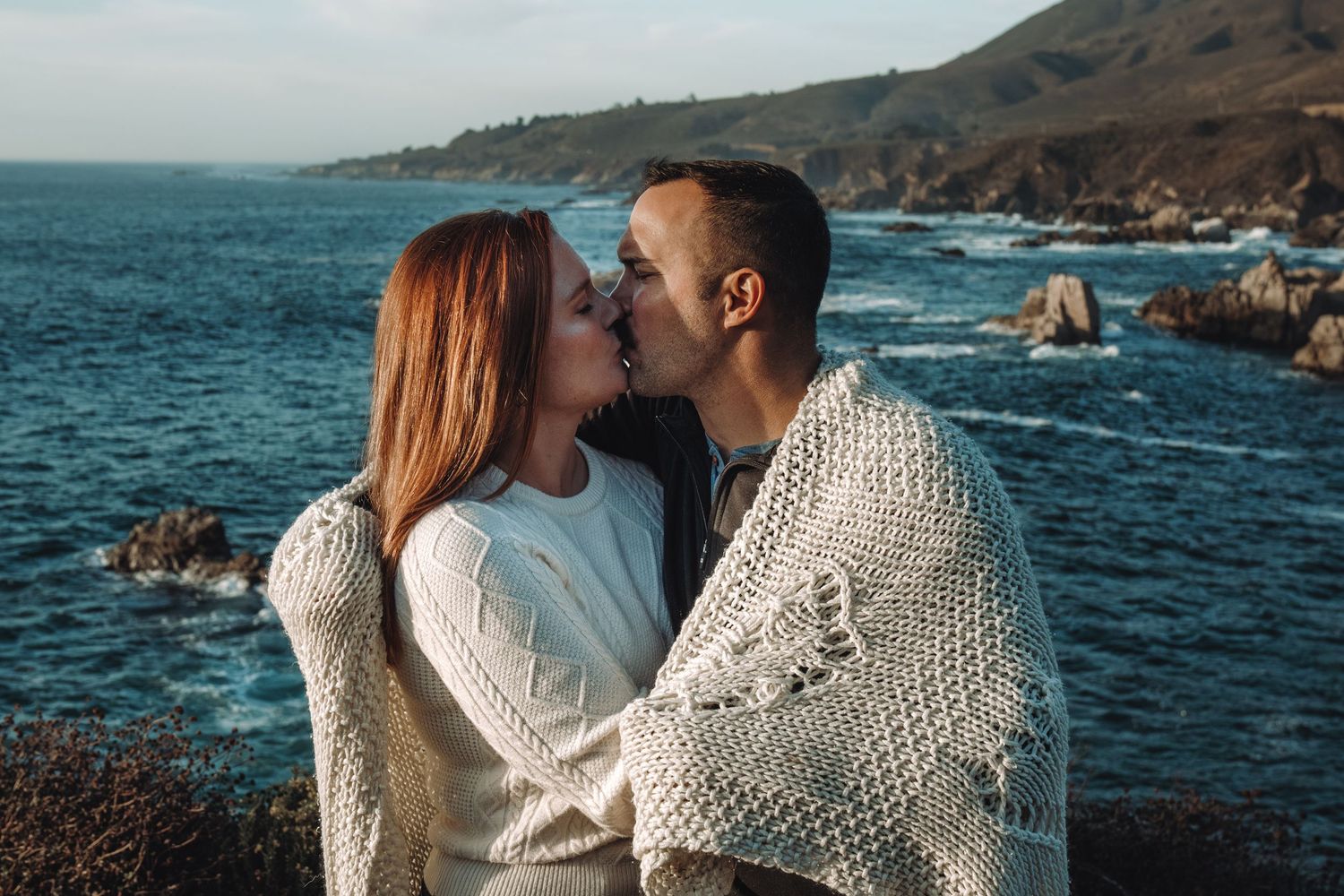 Two people in cozy white sweaters share an intimate moment along a rocky coastline with blue ocean waters and cliffs.