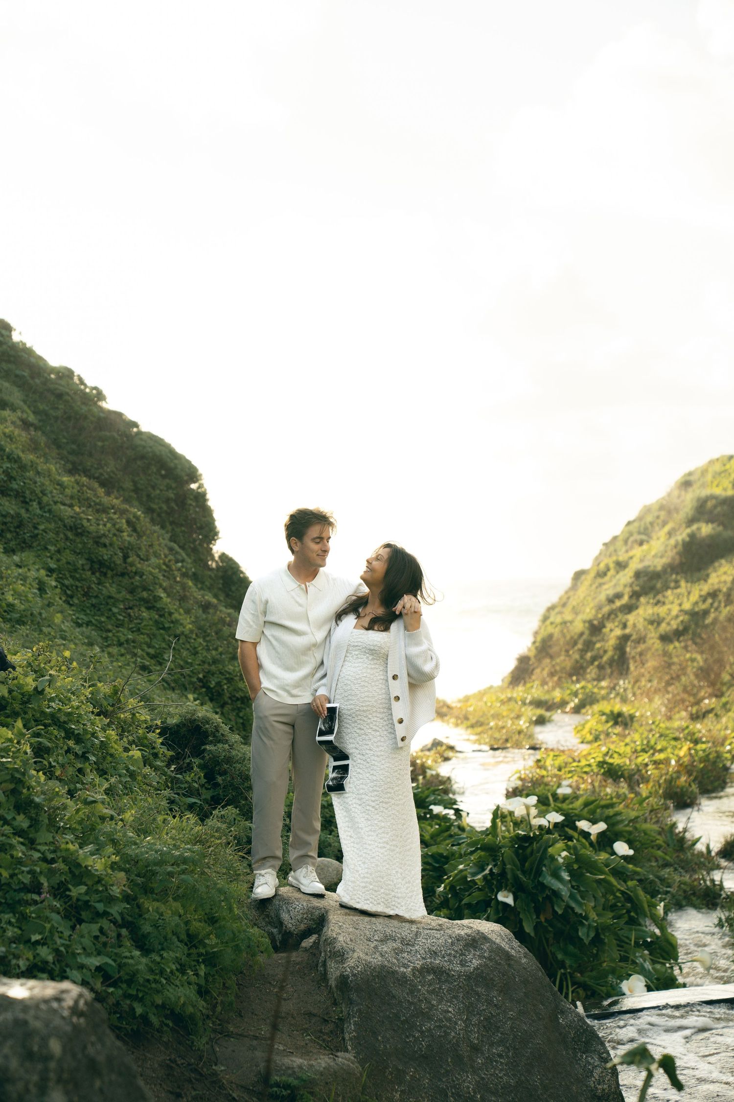 Couple in white outfits standing on rocks surrounded by lush greenery and ocean views.