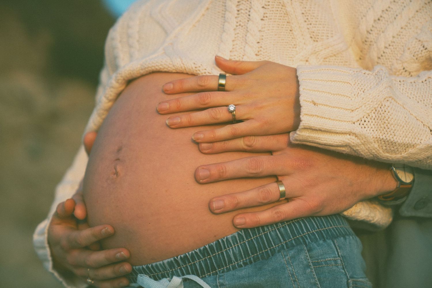 Couple placing hands on pregnant belly, wearing wedding rings, in soft natural light.