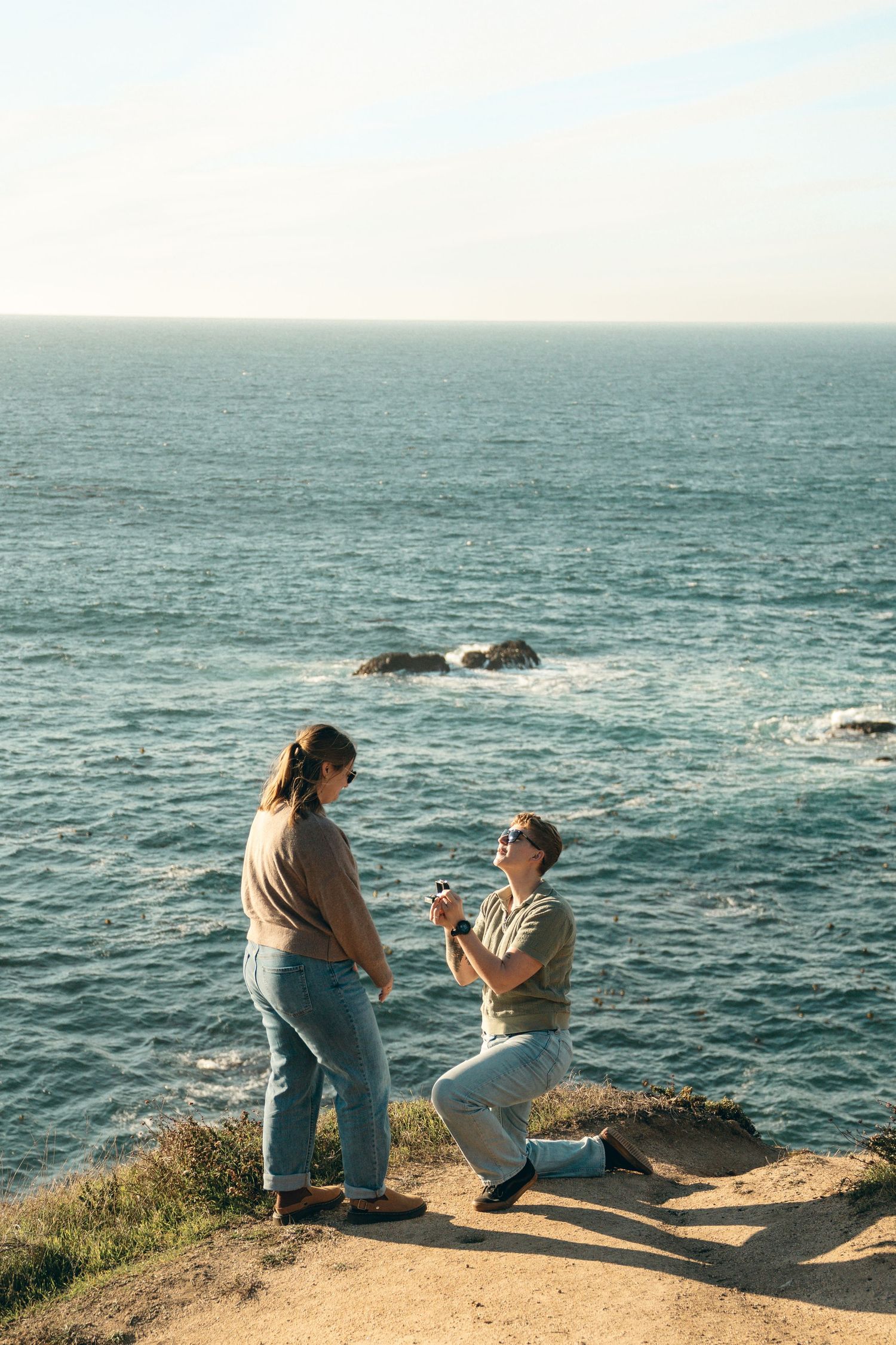 Two people sit at the edge of a coastal cliff overlooking the vast blue ocean on a sunny afternoon.