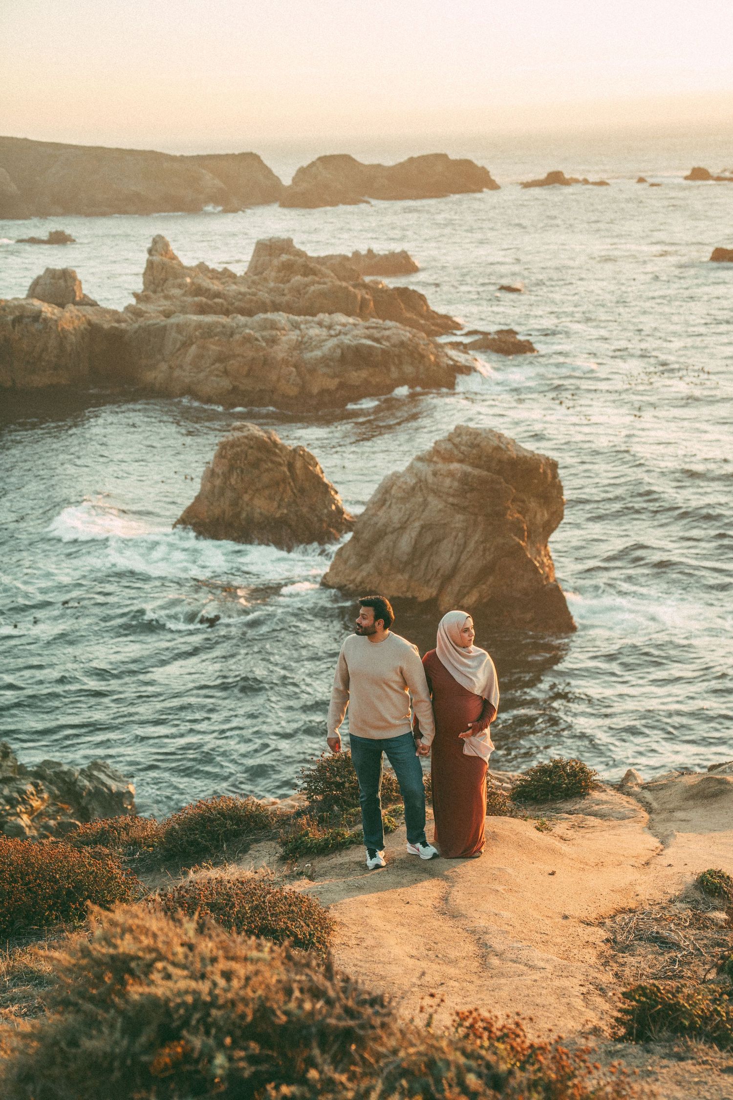 Couple enjoying scenic coastal views from cliff edge overlooking rocky Pacific Ocean shore at sunset.