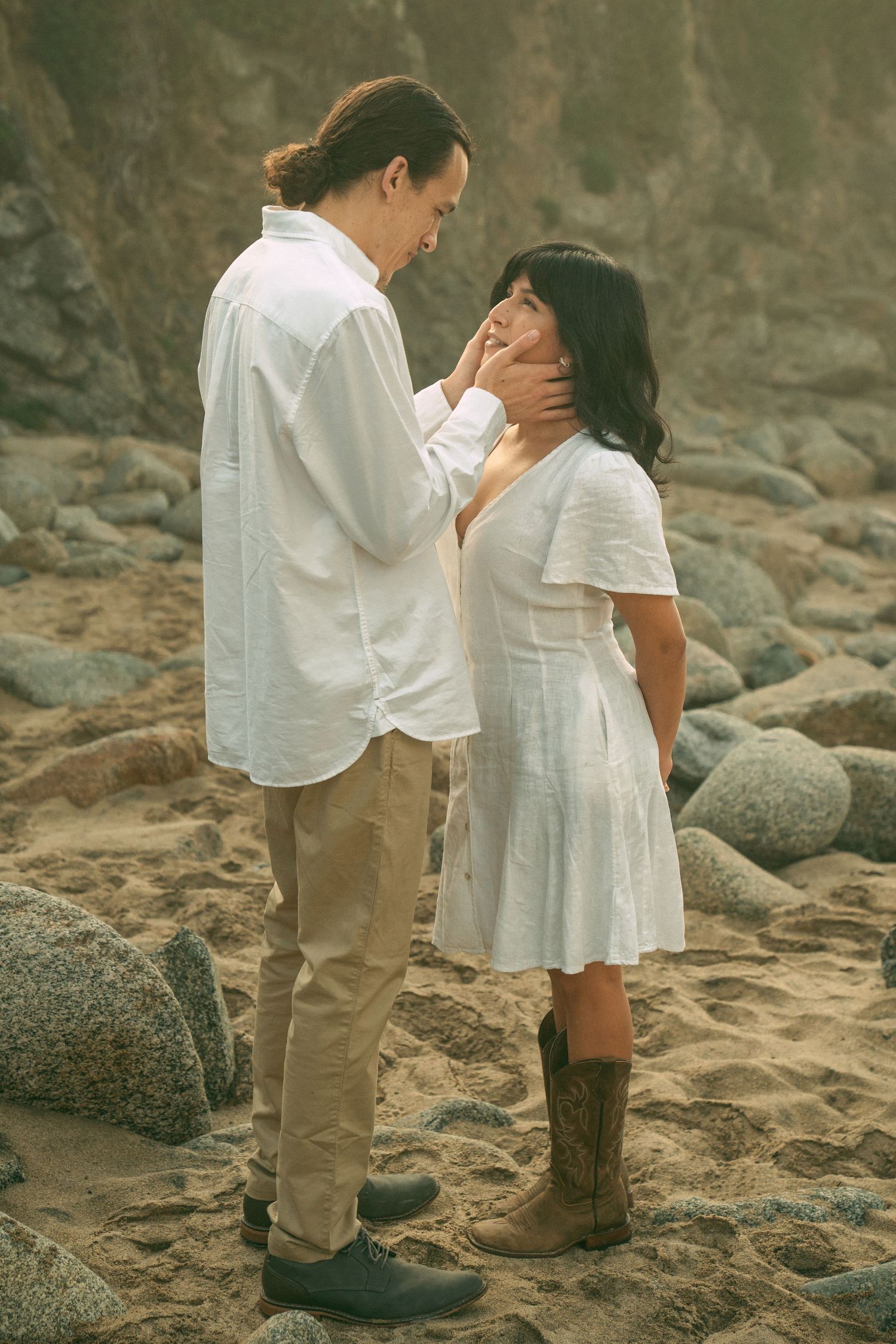 A romantic couple in white outfits sharing tender moments on a rocky beach at sunset.