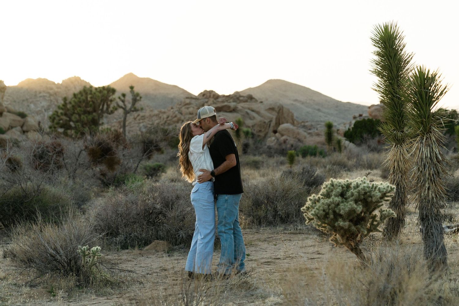 A couple stands among desert plants and Joshua Trees in a golden hour landscape at Joshua Tree National Park.