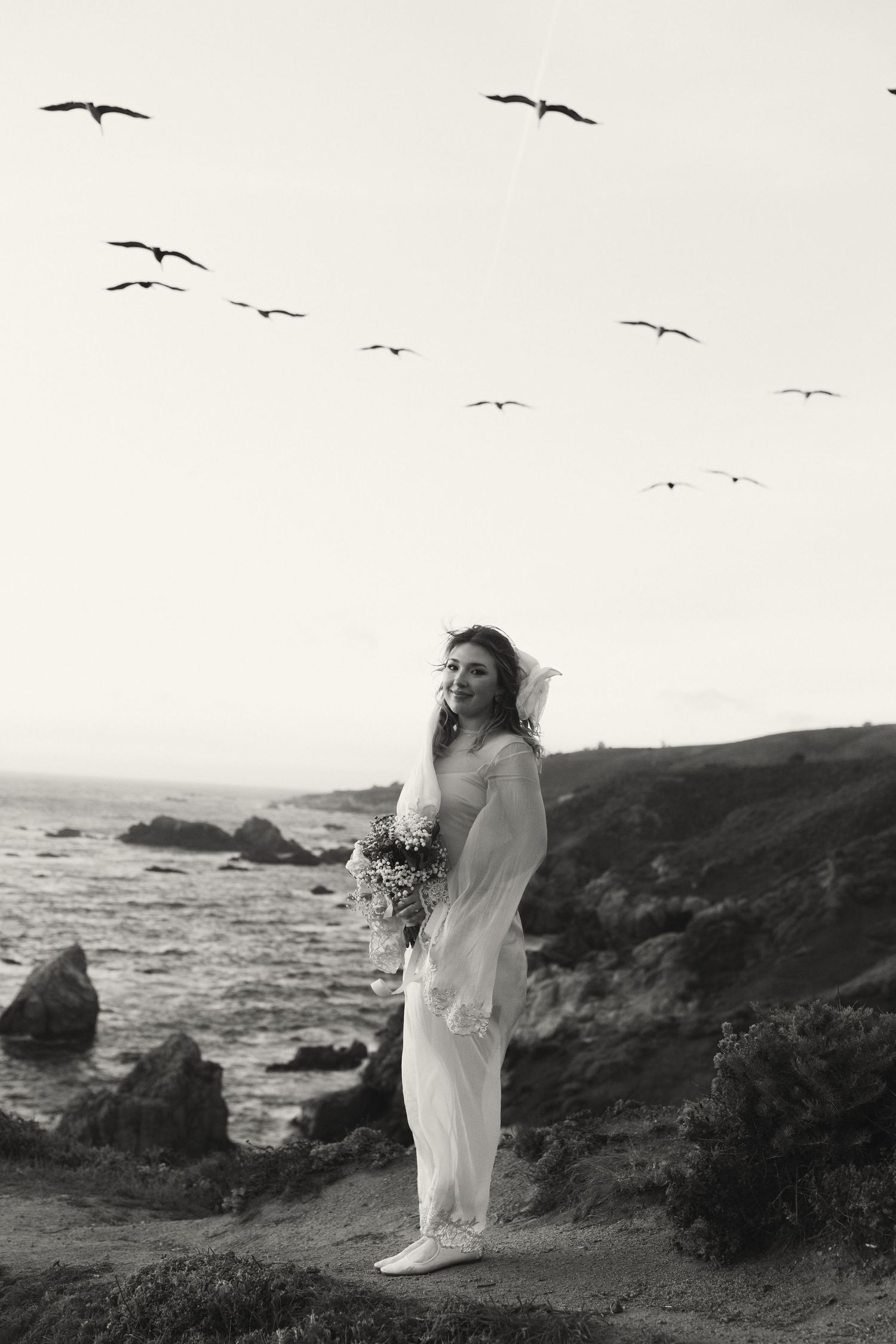 Bride in flowing white dress holding bouquet on coastal cliffside with birds flying overhead in black and white photo.