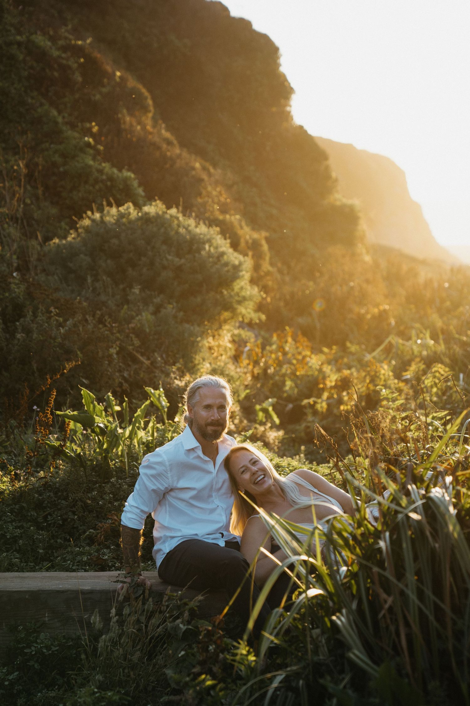 Couple relaxing together in garden at golden hour with sunlight streaming through trees and foliage.