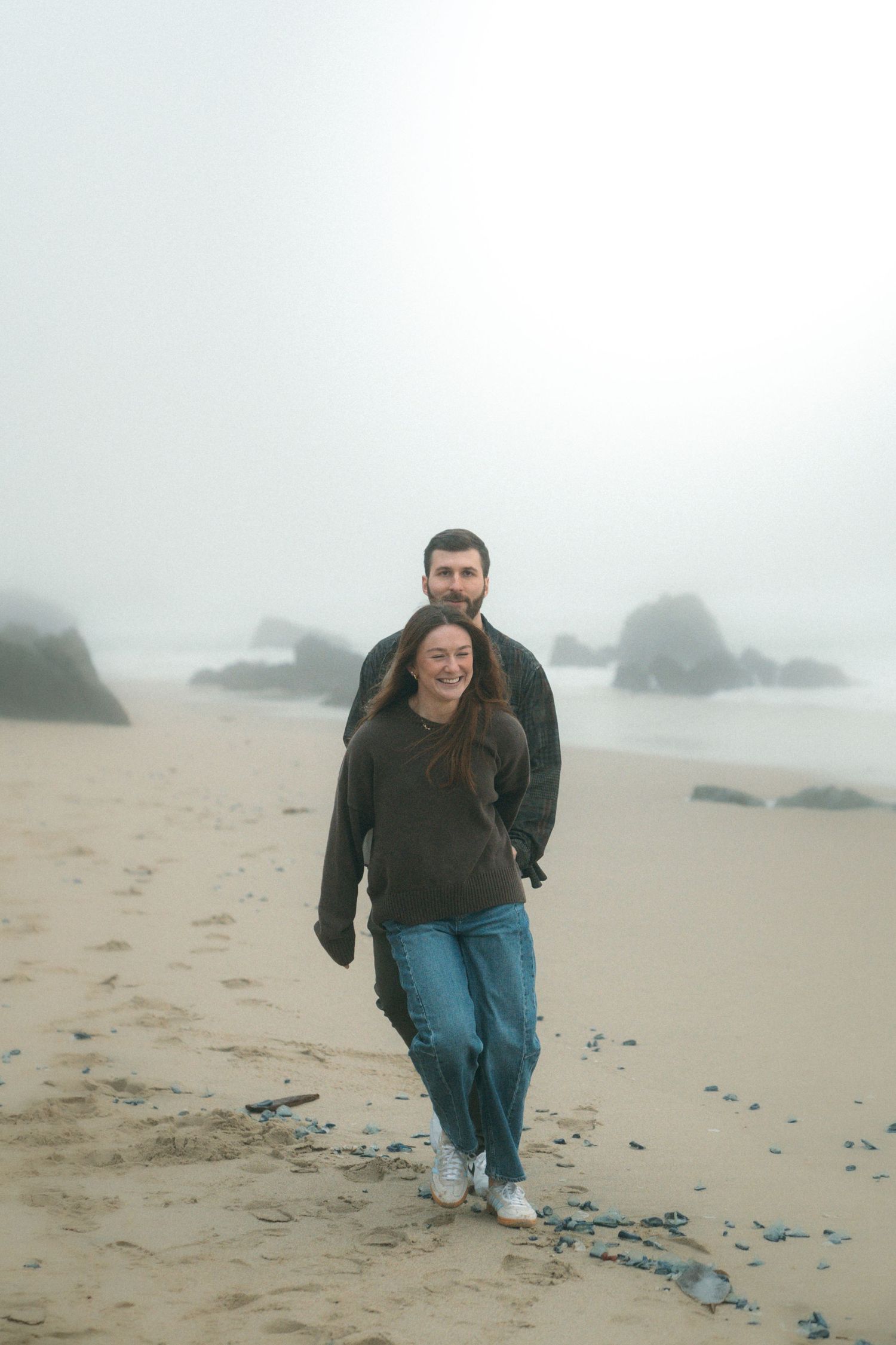 Two people walking together on a foggy beach with large rocks visible in the distance.