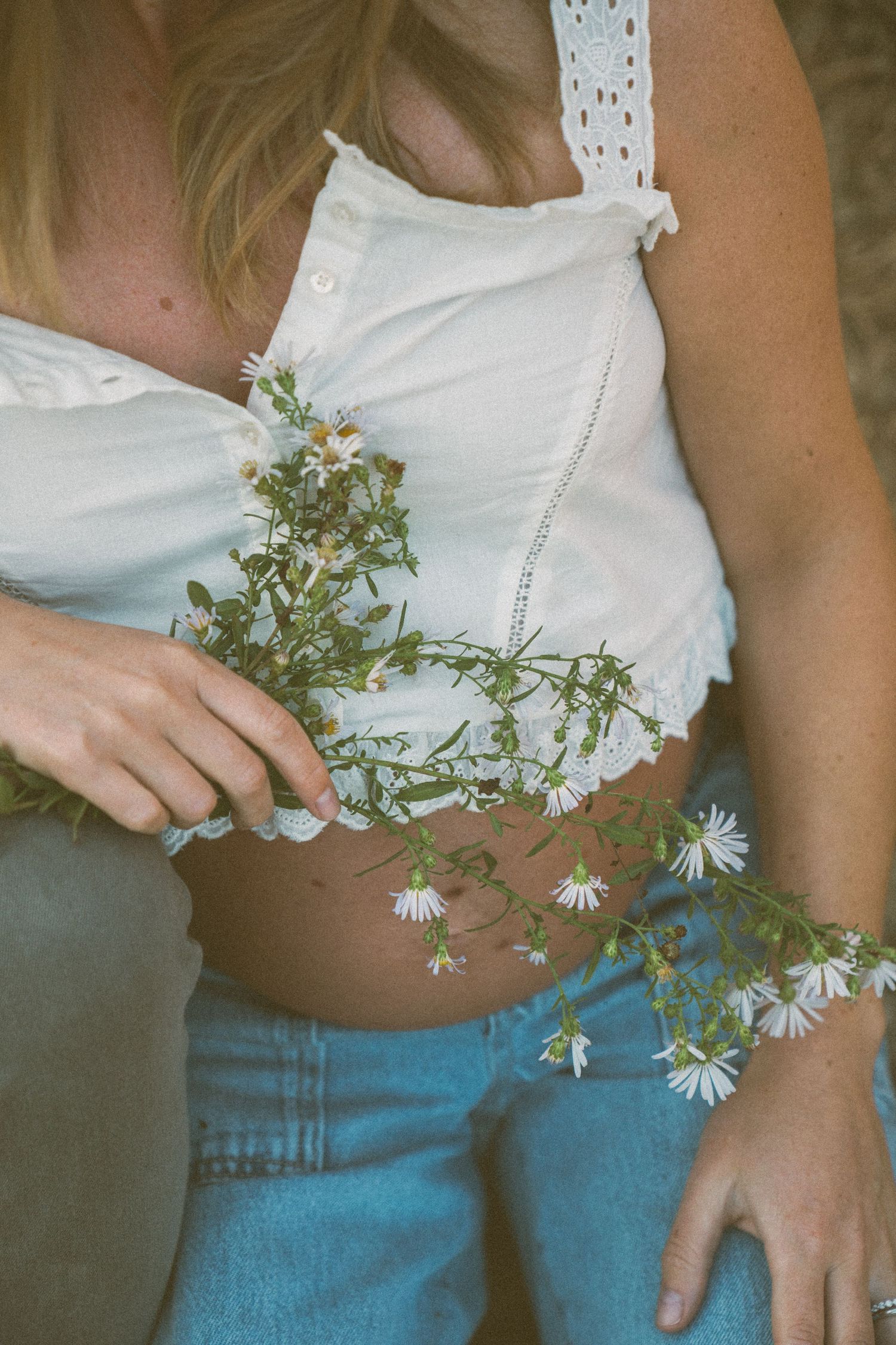 A person in a white crop top and blue jeans holds delicate white daisies against their torso in a vintage-style photo series.
