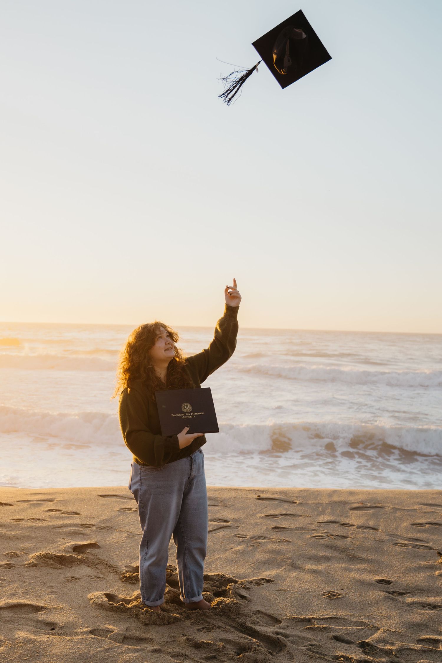 Graduate celebrates on beach at sunset by throwing graduation cap in the air while sitting in the sand.