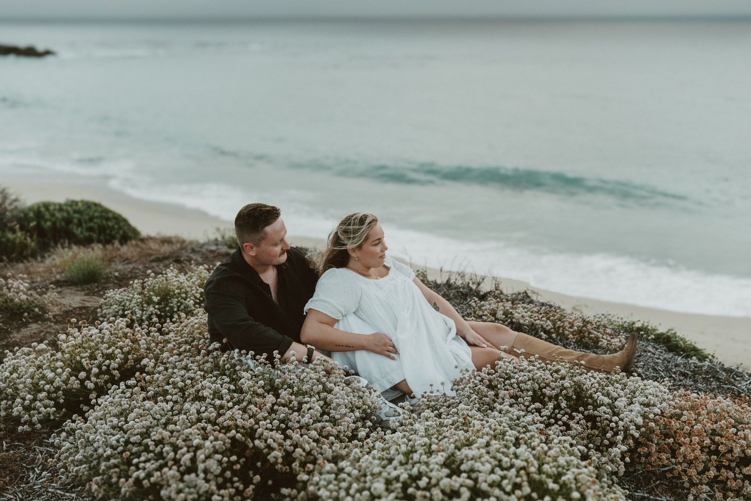 Couple in wedding attire embraces on coastal cliffs surrounded by white wildflowers with ocean views.