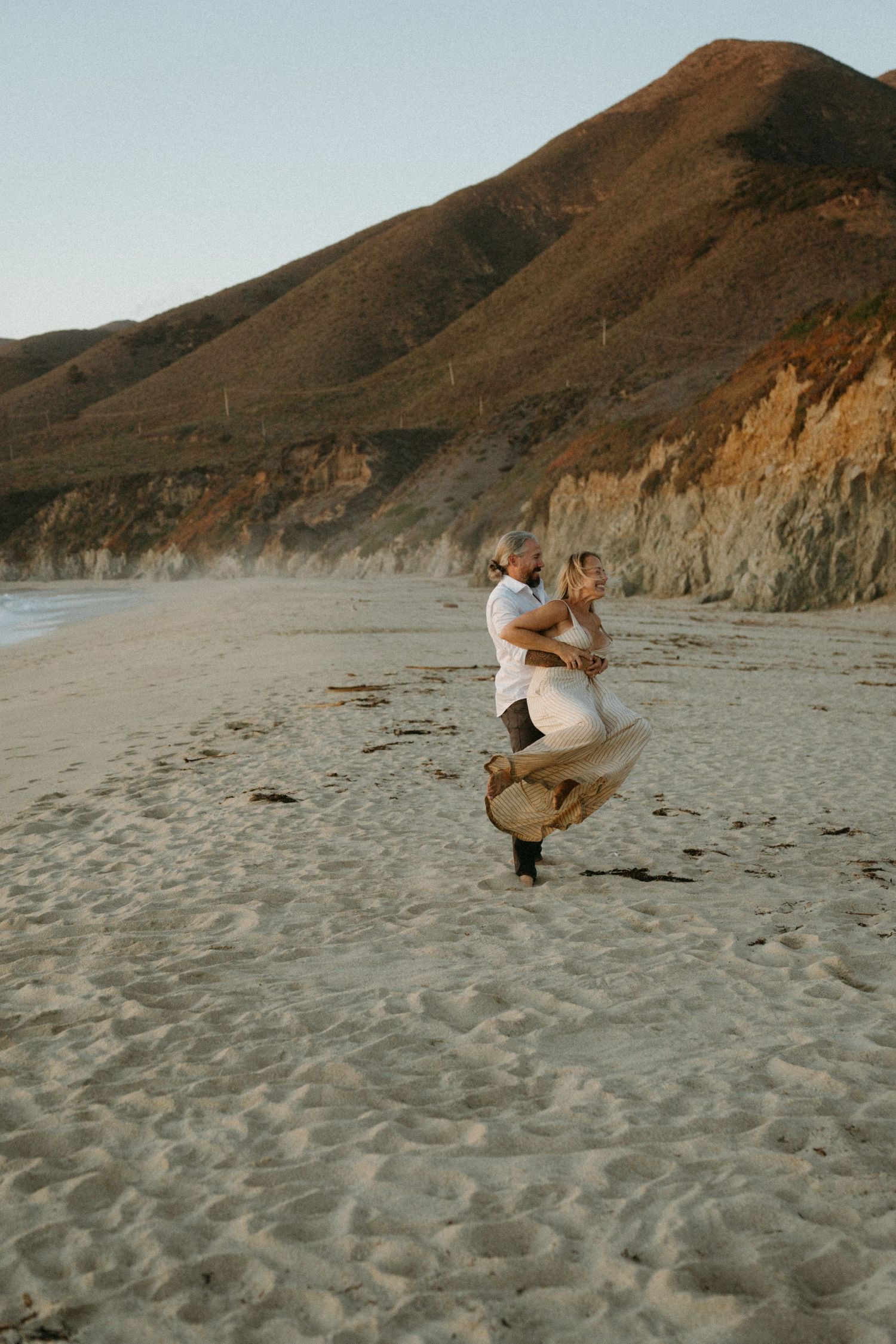 Two people playfully dance together on a sandy beach with dramatic coastal cliffs in the background at sunset.