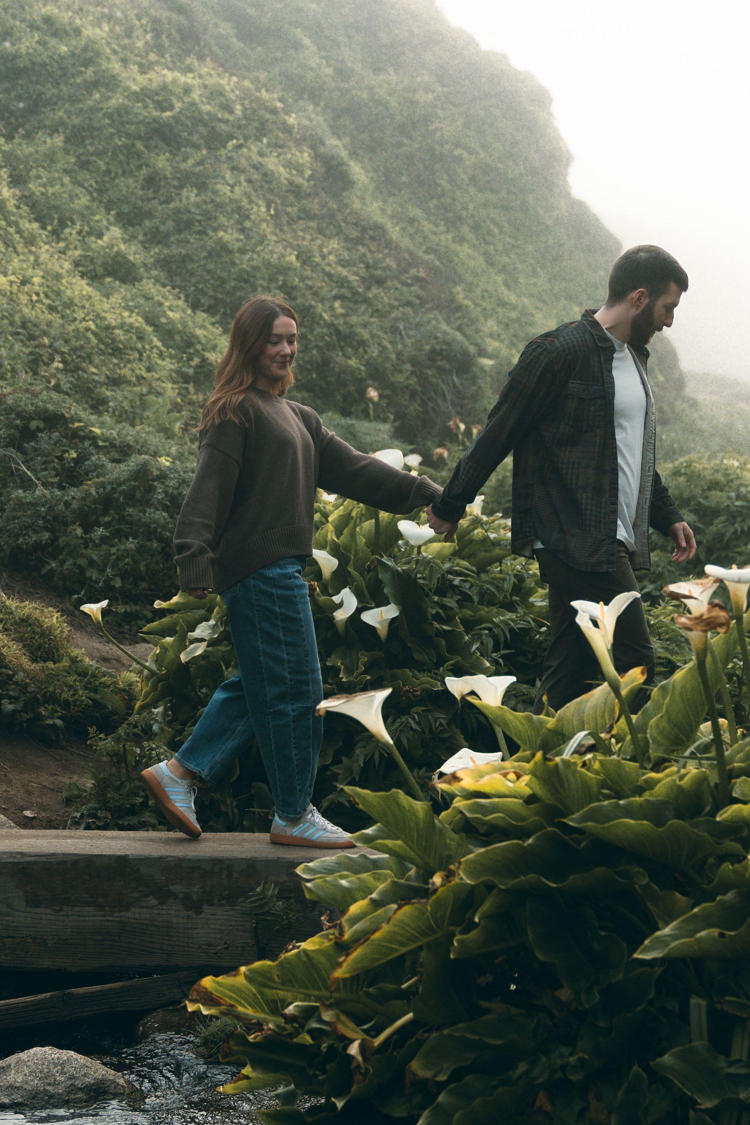 A couple walking hand in hand along a mountain path with lush vegetation.