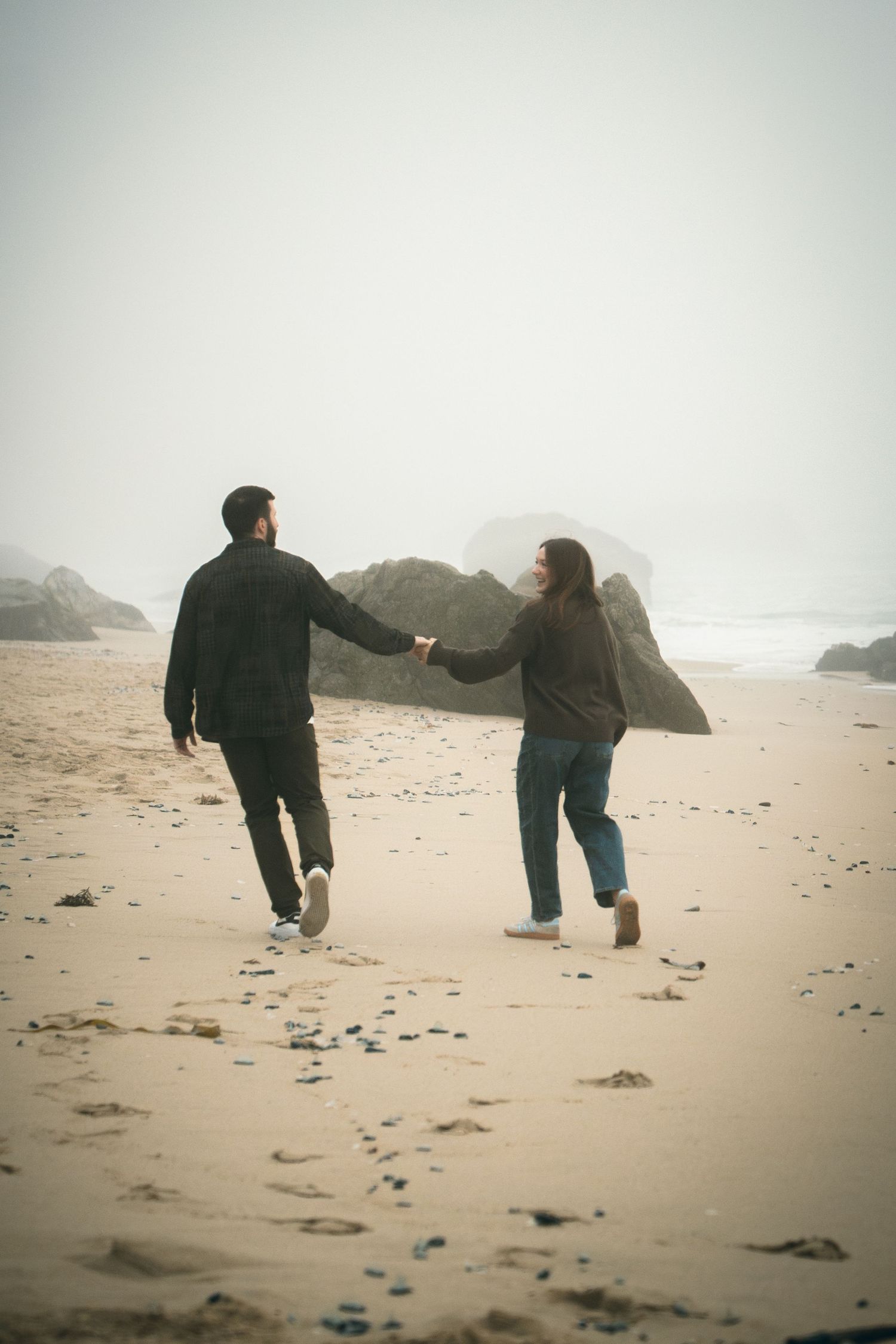 Two people holding hands while running along a misty beach with rocks in the background.