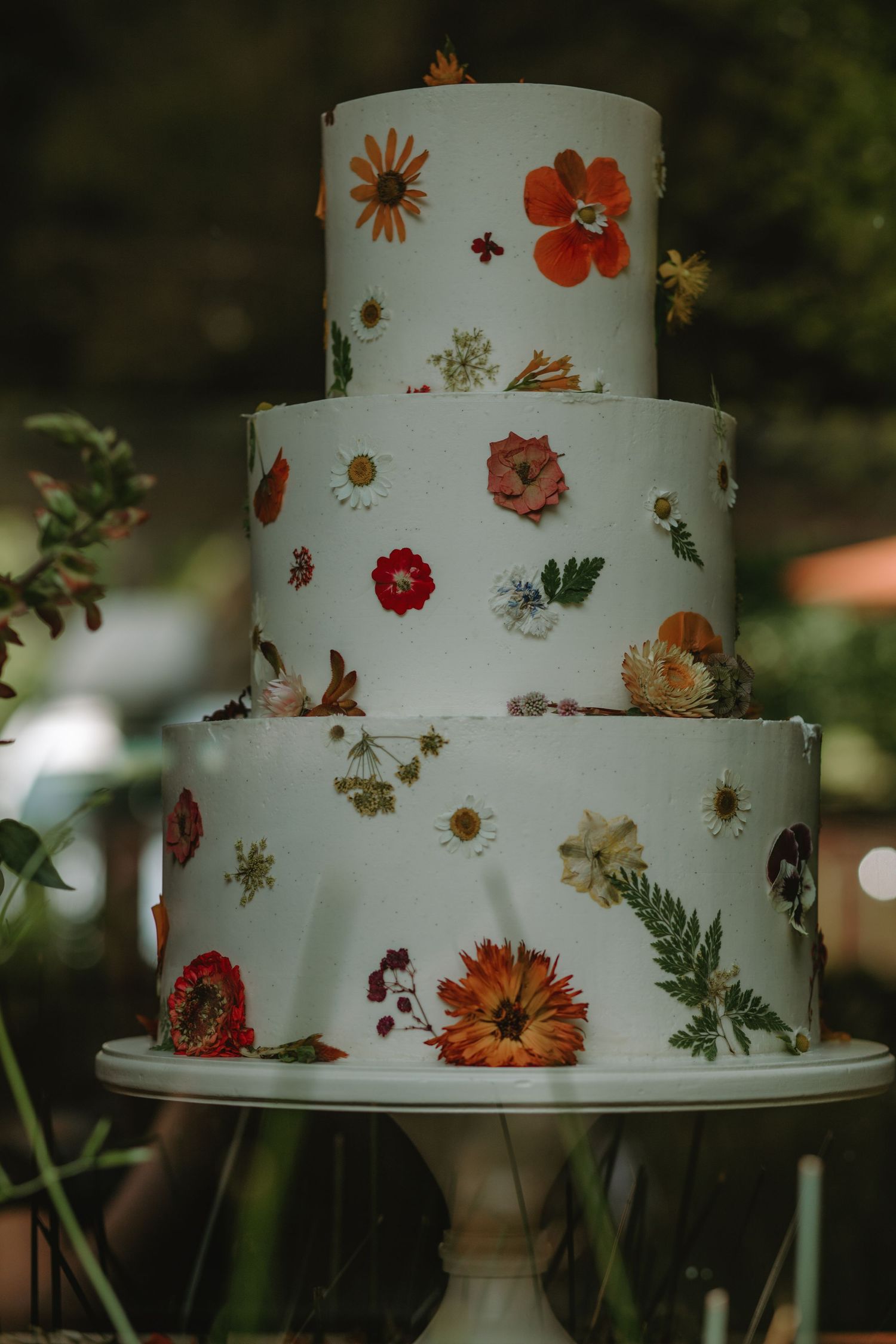 Three elegant white wedding cakes decorated with pressed edible flowers in orange, red and pink displayed on cake stands.