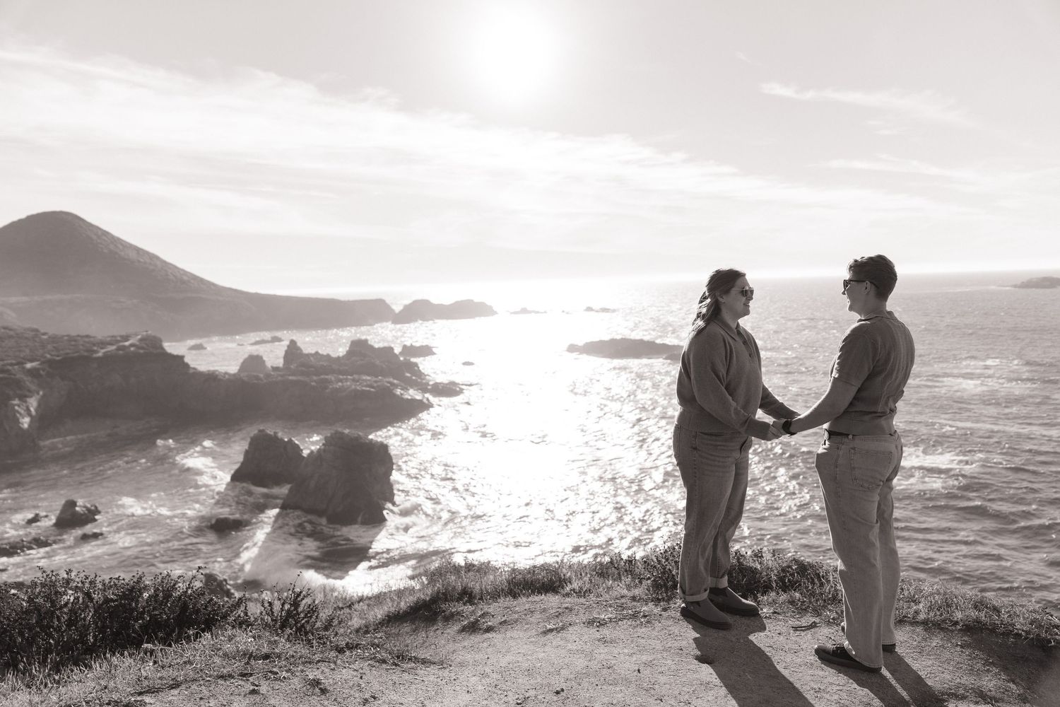 Silhouetted figures stand along a scenic coastal cliff overlooking the Pacific Ocean at sunset.