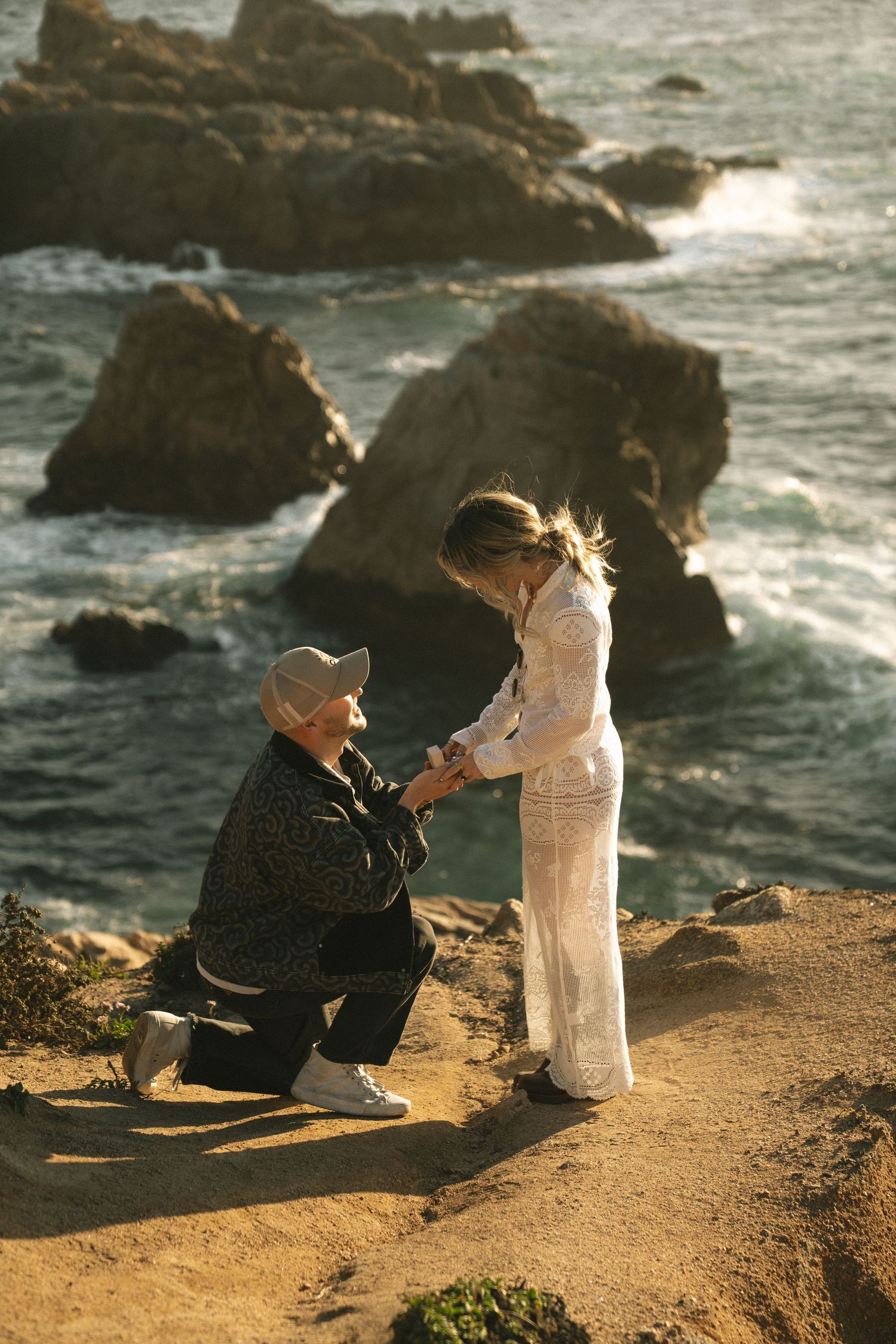 Person proposing on one knee to partner on rocky coastal cliff with ocean waves and rock formations in background.