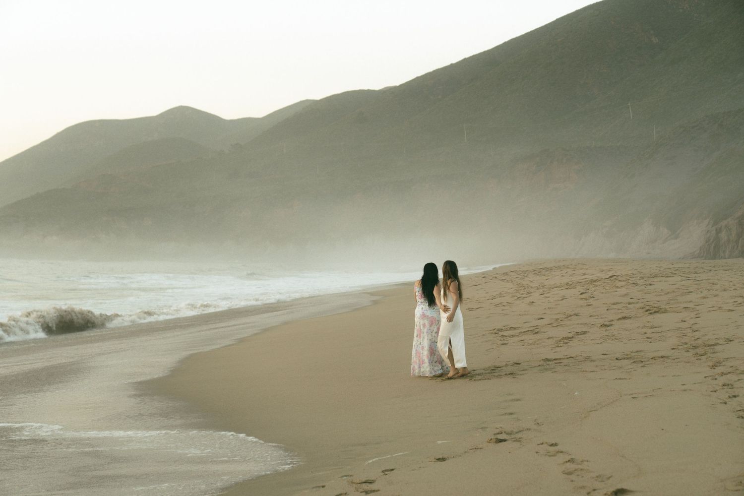Two women in white dresses walking along a misty beach with rolling waves and scenic mountain backdrop.