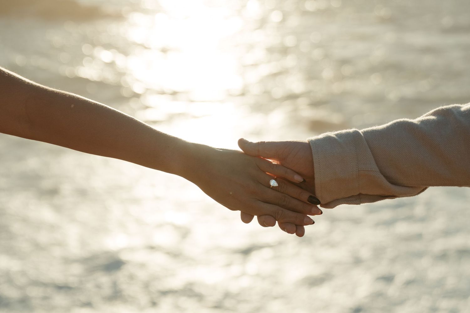 Two hands join together in a tender moment against a glowing sunset backdrop over the water.