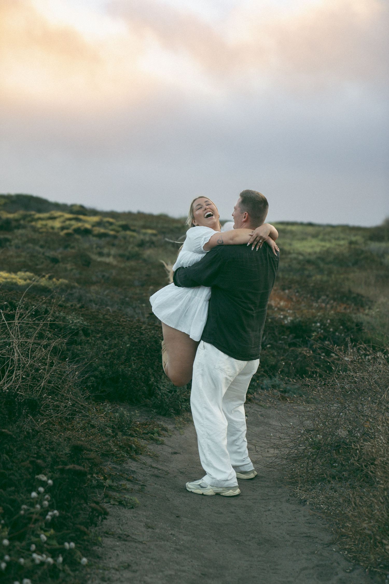 Two people in contrasting black and white outfits dance playfully on a hillside trail at dusk.