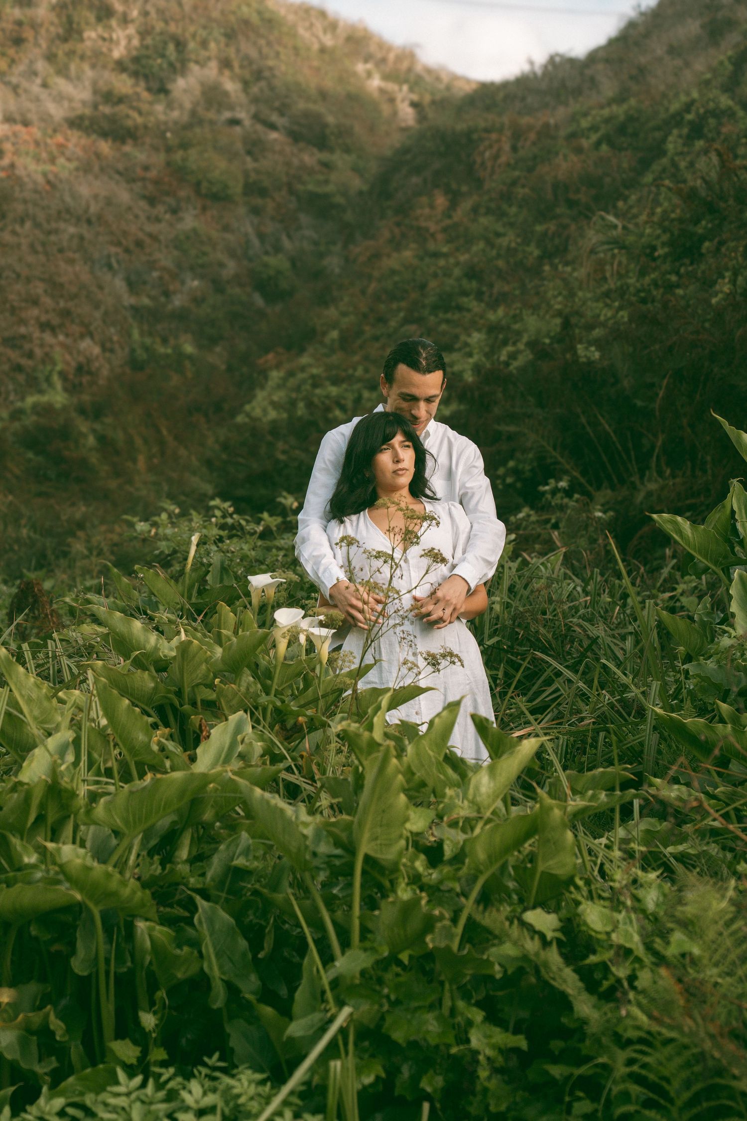 Two people sit among lush green foliage in a mountainous landscape during golden hour.