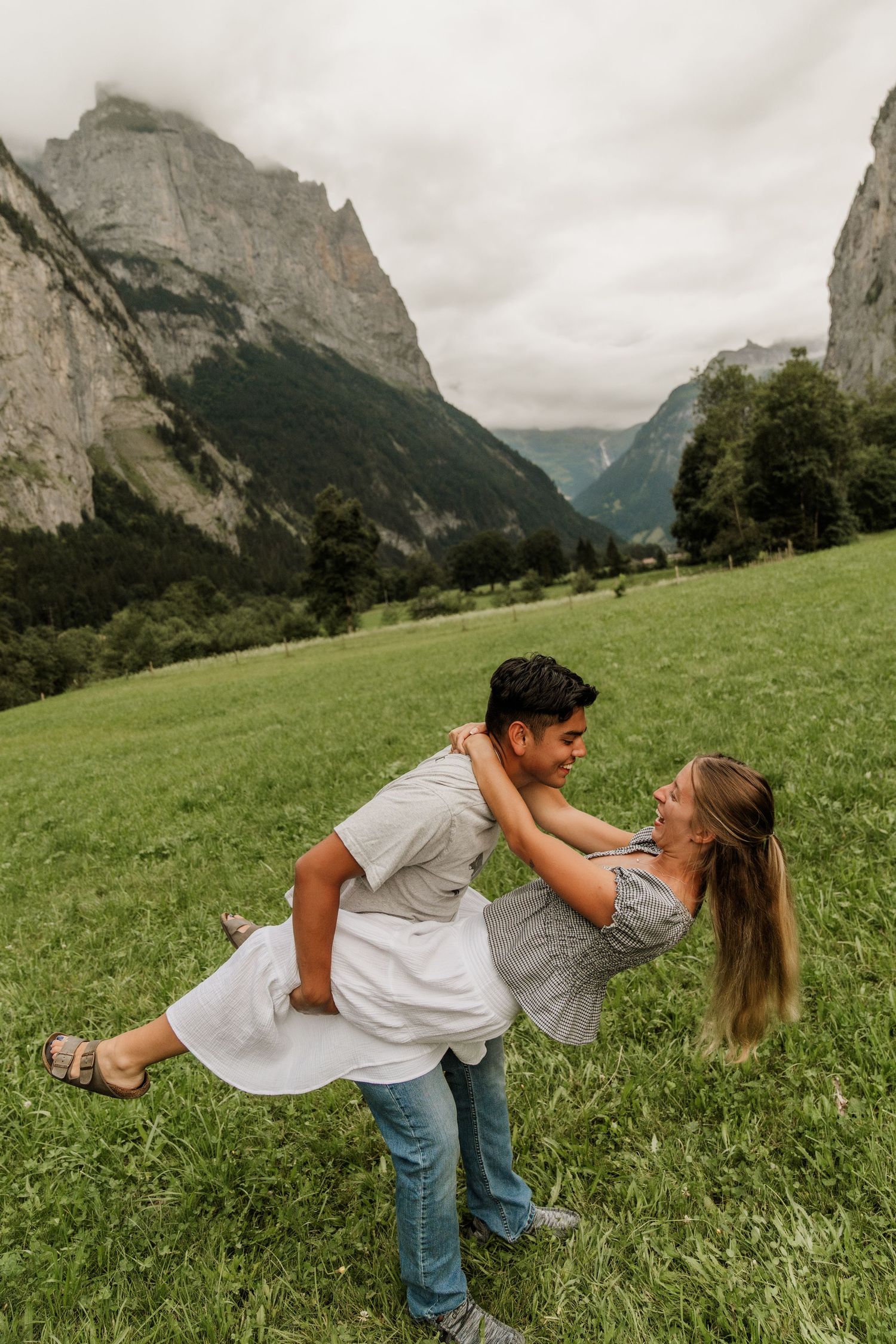 A couple dances playfully in a grassy meadow with dramatic mountain peaks and valleys in the Swiss Alps background.