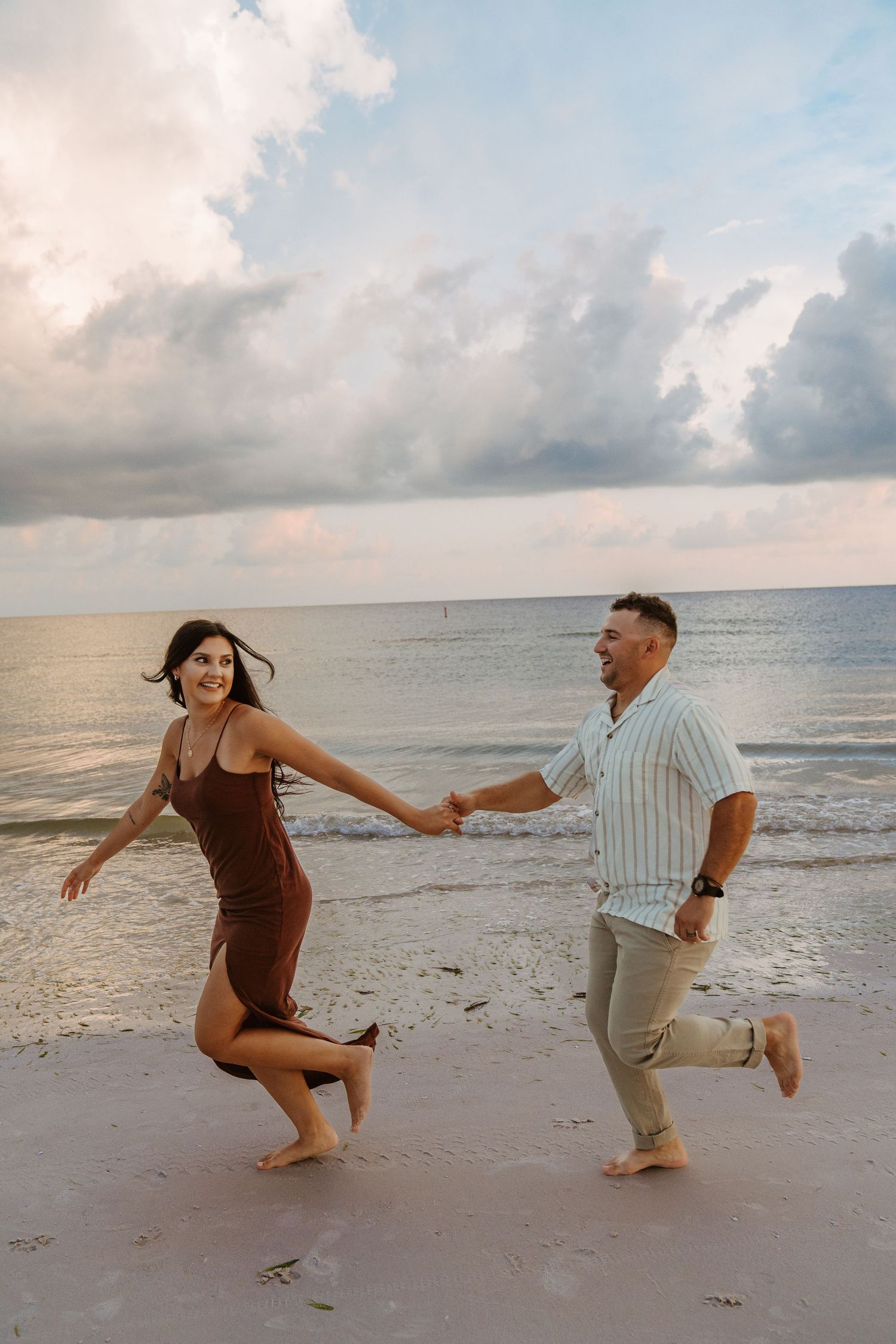 A playful couple runs along the shoreline at sunset holding hands.
