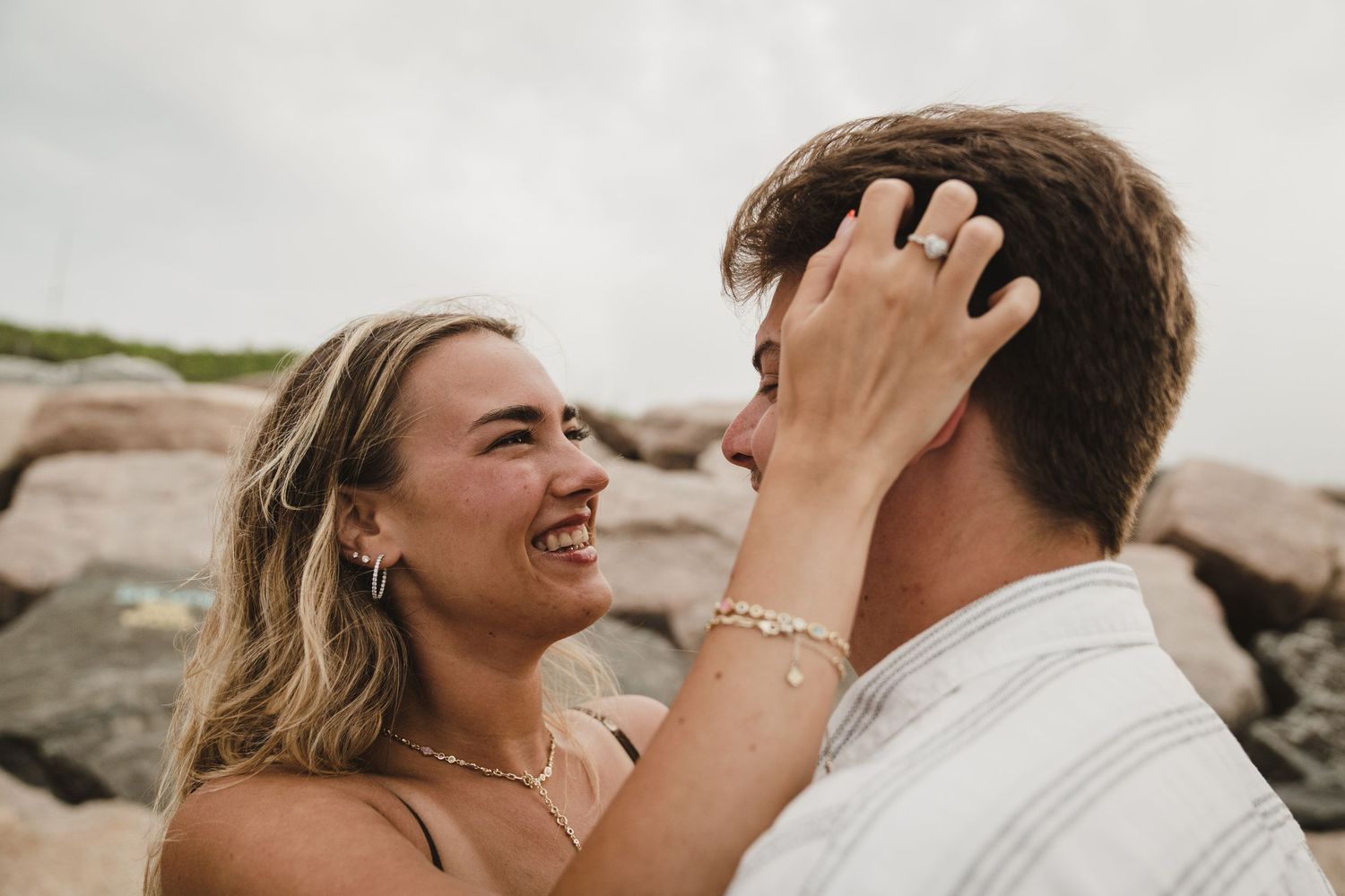A joyful couple embraces and laughs together during a romantic photoshoot on a rocky beach at sunset.