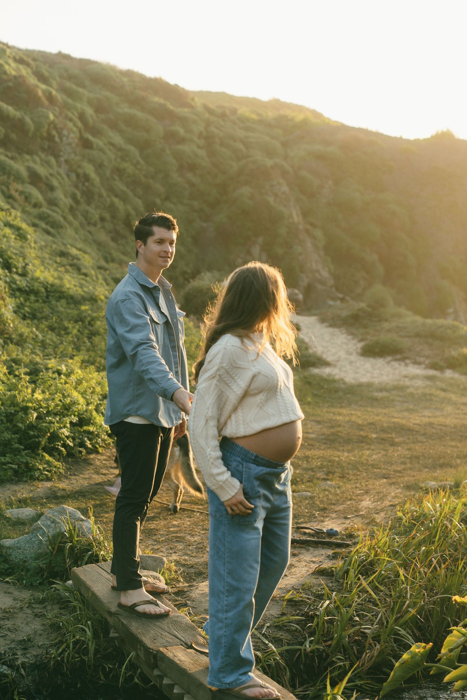 Pregnant couple holding hands at sunset on a scenic coastal hillside path.