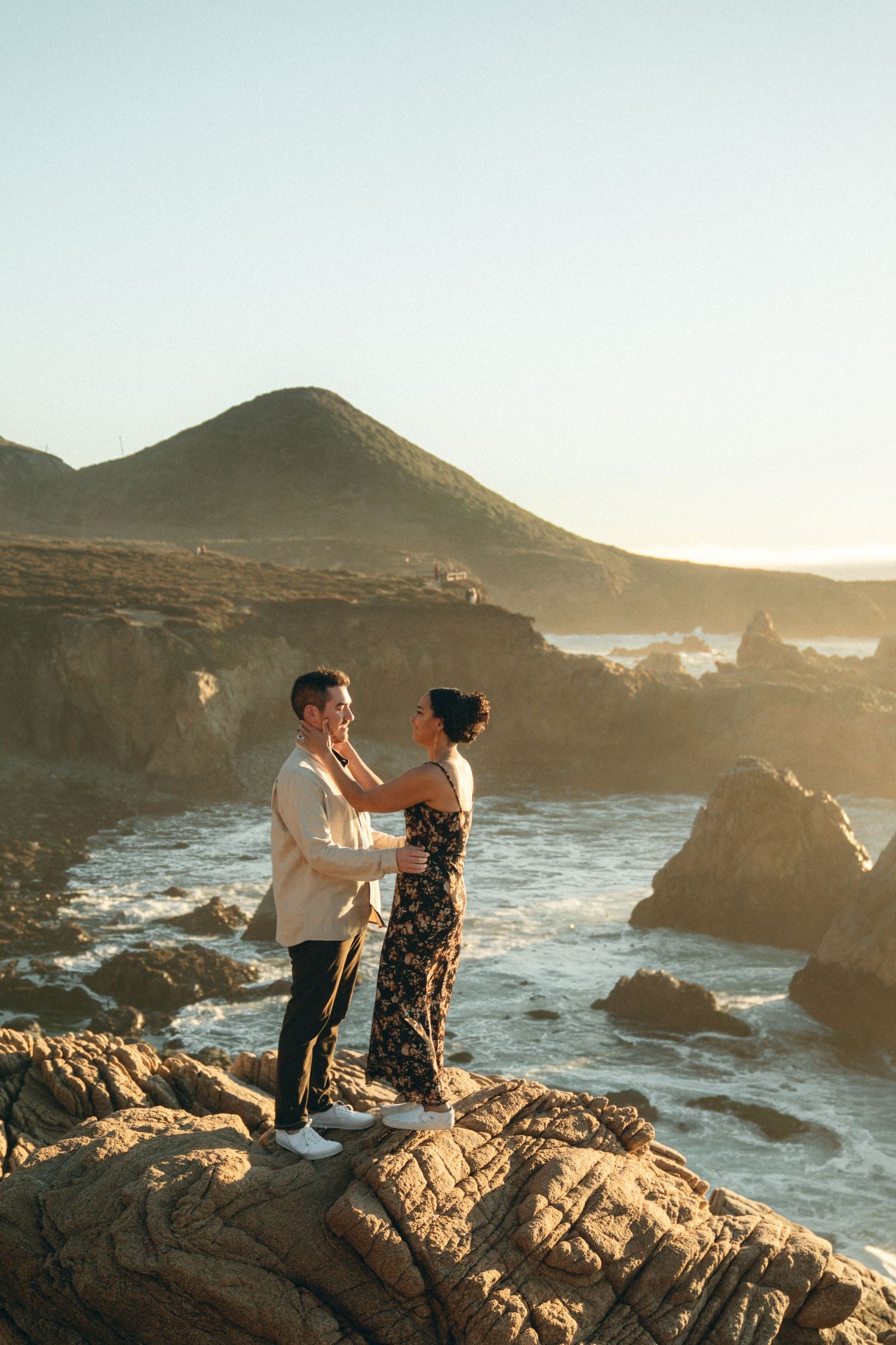 Couple sitting together on coastal rocks at sunset while ocean waves crash against the rugged shoreline.