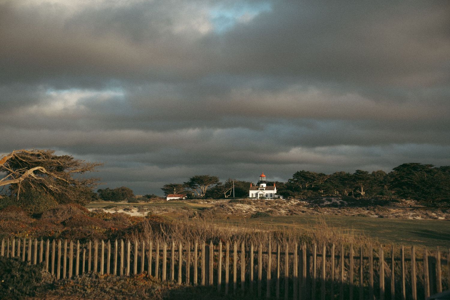 Dramatic cloudy sky over coastal landscape with wooden fence and buildings in the distance.
