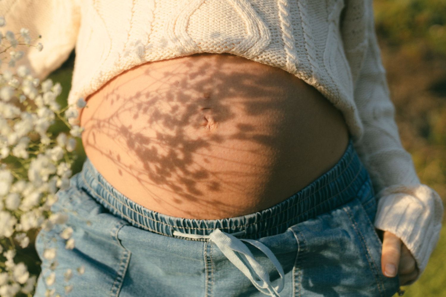 Close-up of a pregnant belly with stretch marks, wearing a white crop top and blue jeans outdoors.