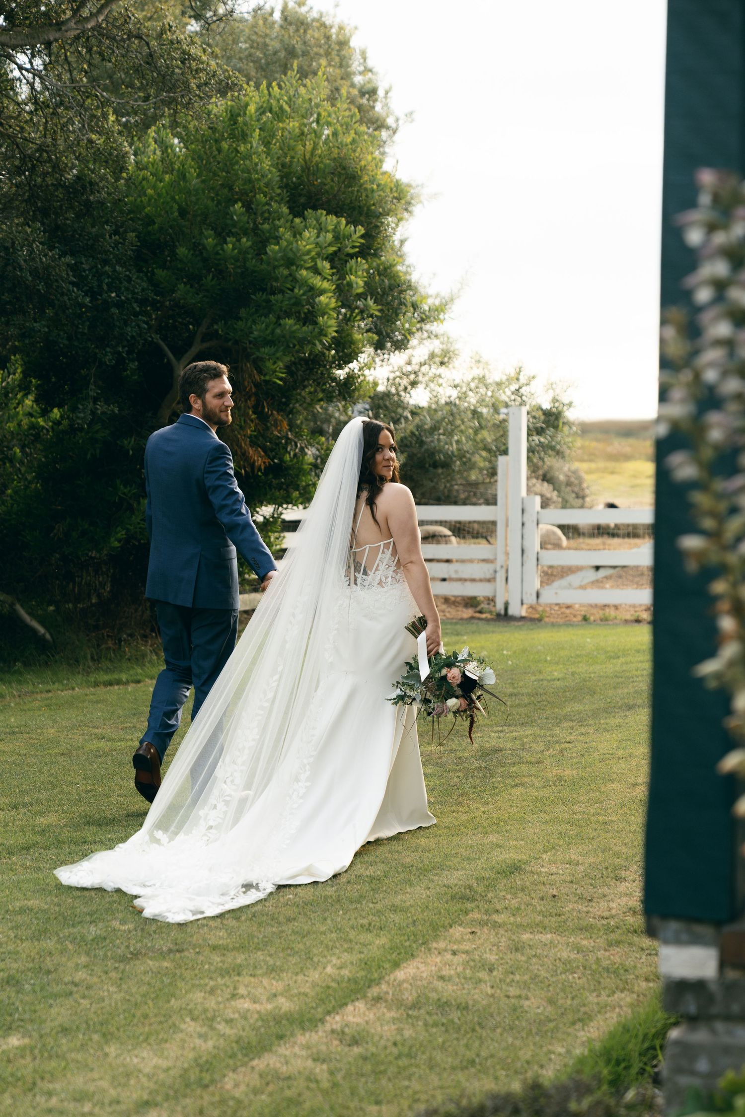 A bride and groom share a romantic moment on a grassy lawn with a rustic white fence in the background.