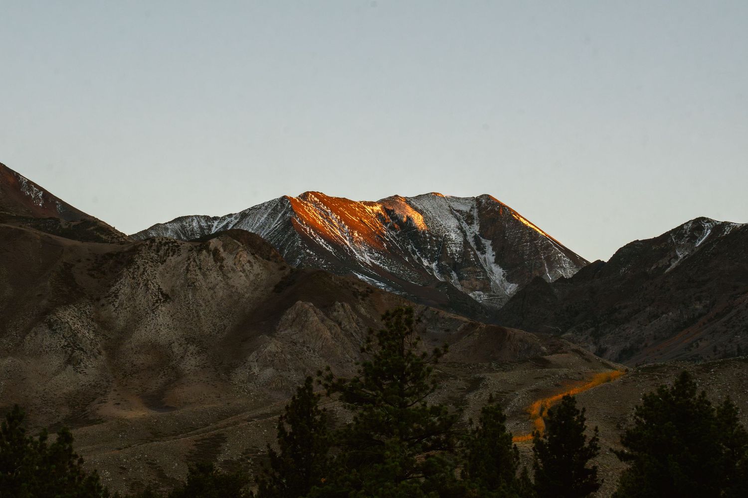 Snow-capped mountain peaks glow orange at sunset against a dark rugged mountain landscape.