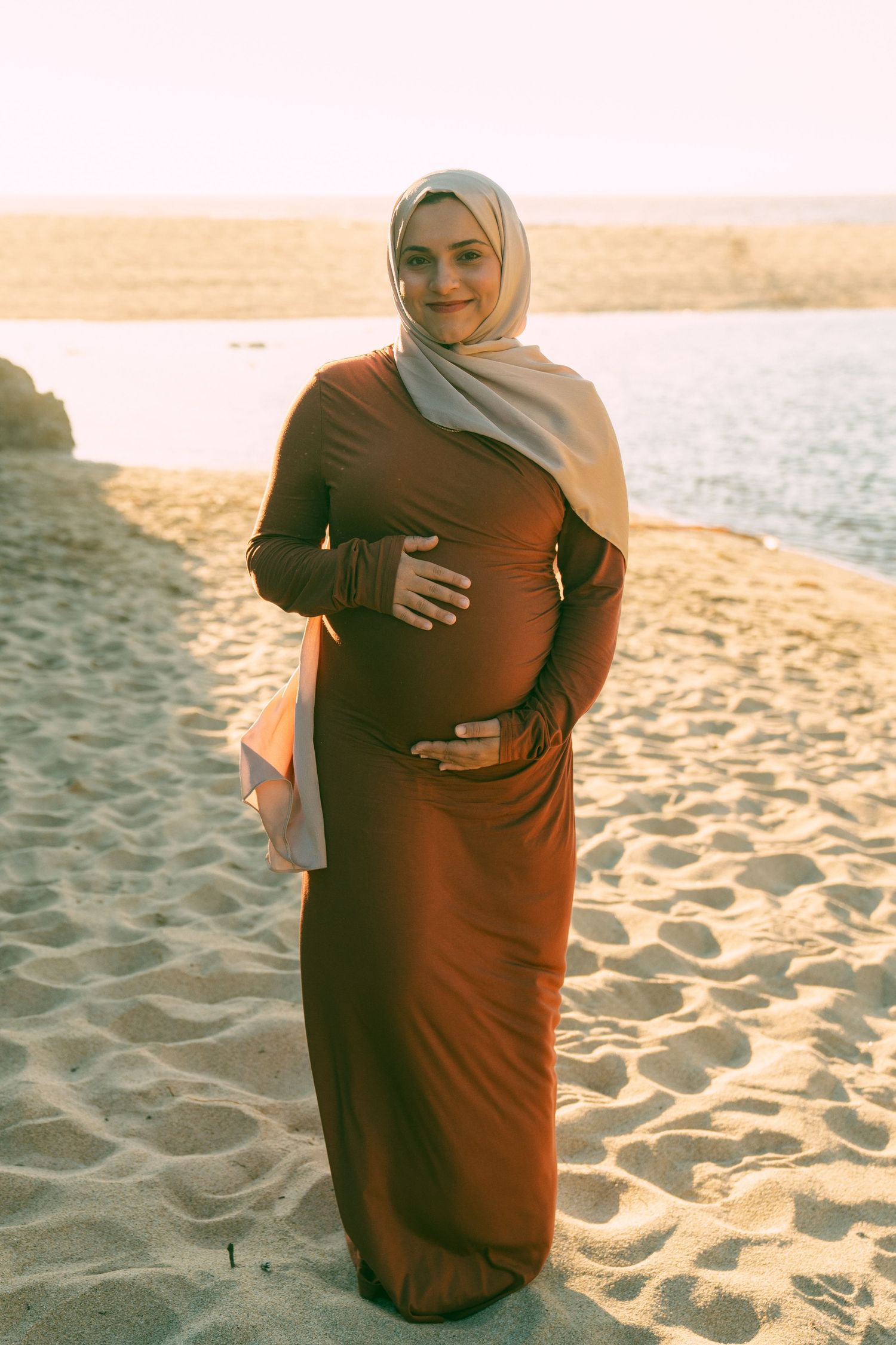 Pregnant woman in brown dress and light hijab poses on sandy beach at sunset, cradling her baby bump.