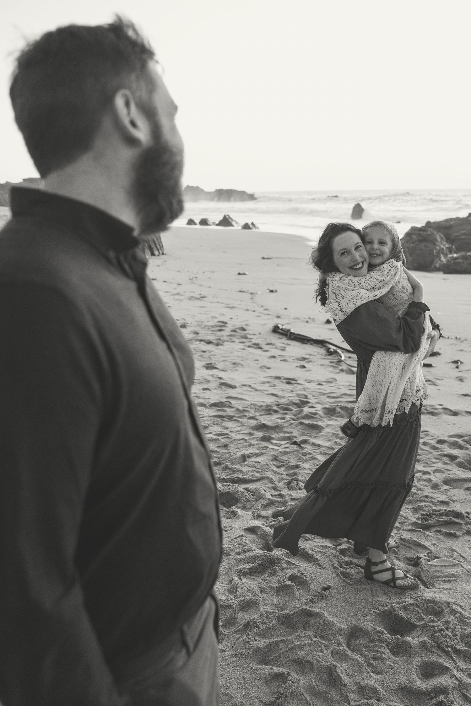 A black and white photo of a family enjoying a moment together on a sandy beach with rocky outcrops.