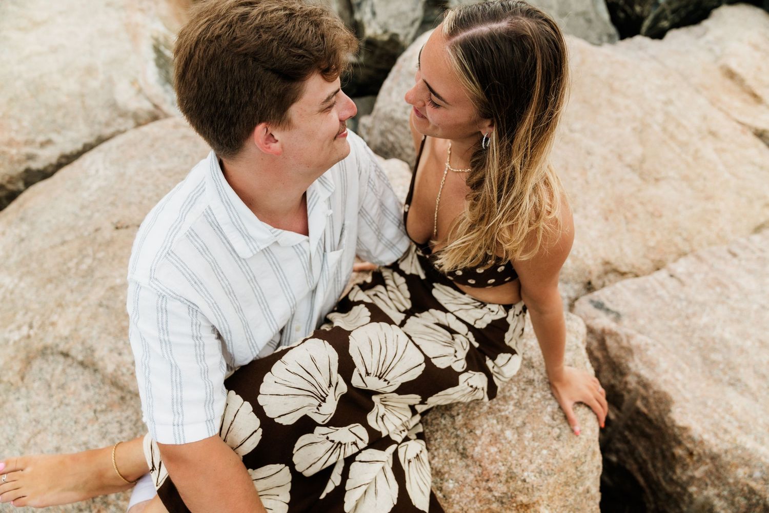A couple dressed in white and floral print share a romantic moment while sitting on coastal rocks at sunset.