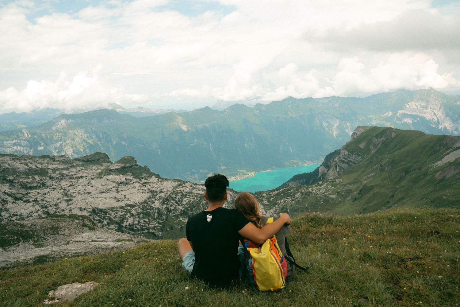 Two people sit together on a mountain summit overlooking a turquoise alpine lake surrounded by rugged peaks.