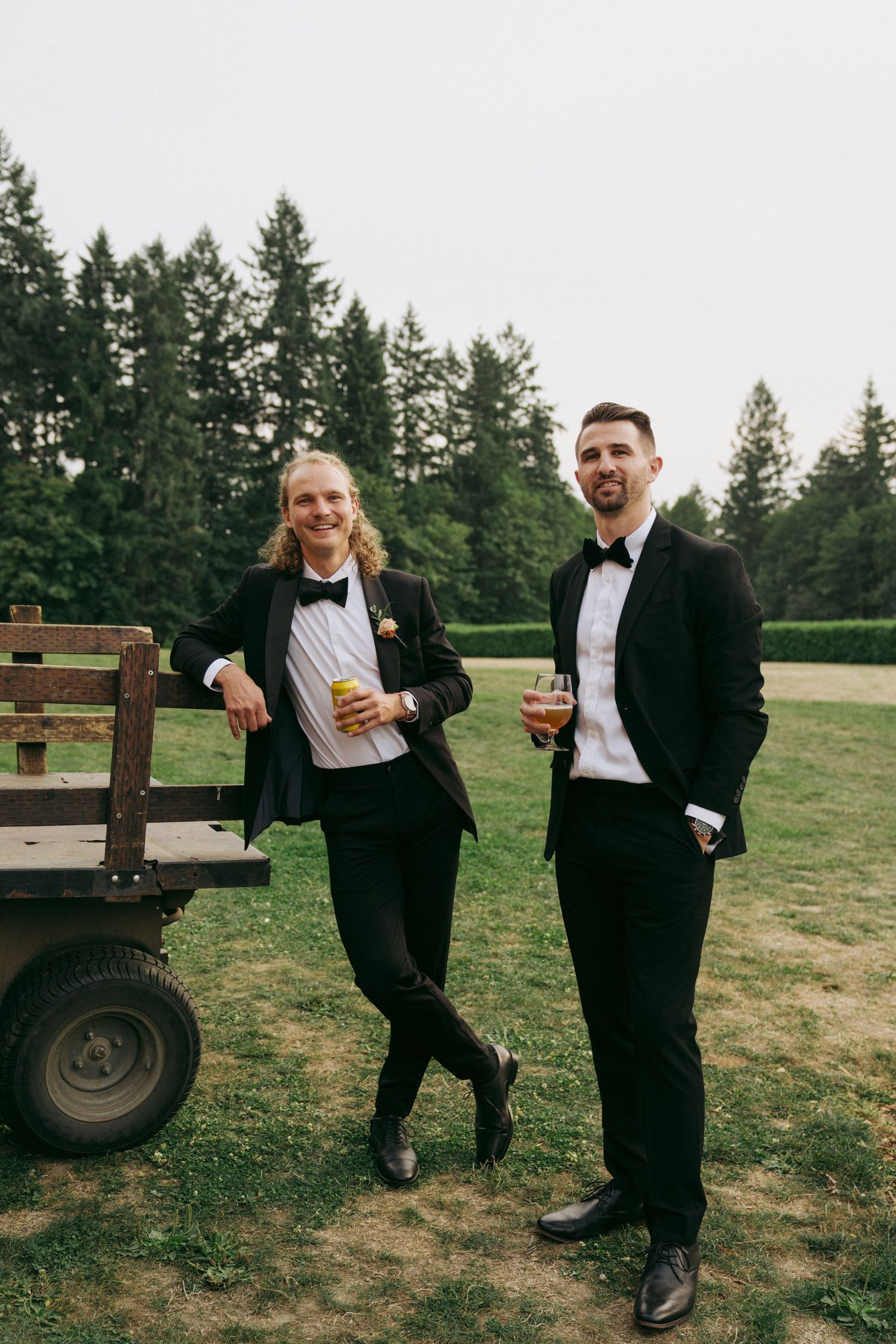Two wedding guests in formal black attire stand casually by a rustic wagon in an outdoor setting.