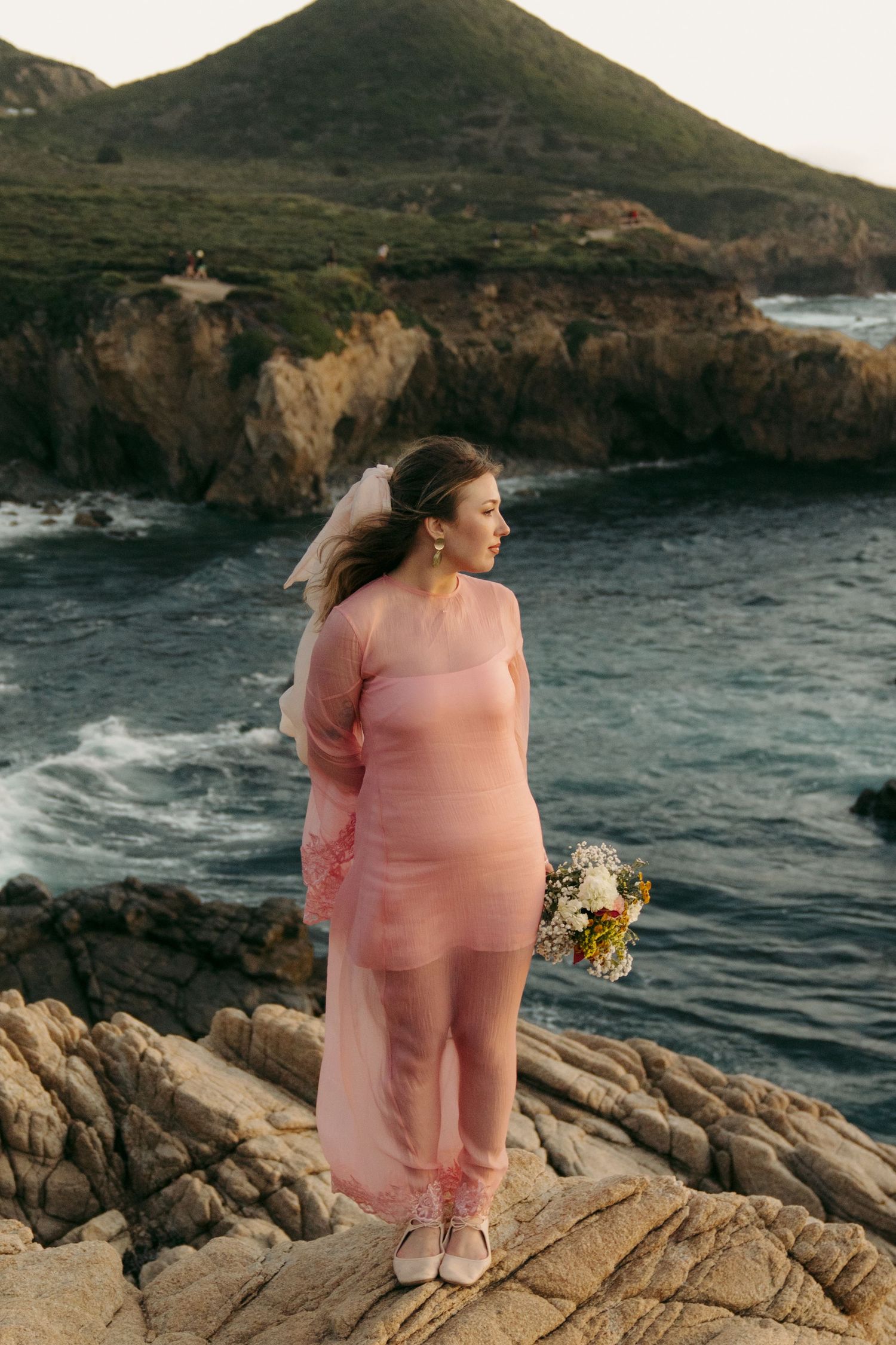 Bride in pink sheer dress stands on rocky cliffside overlooking ocean waves holding floral bouquet.