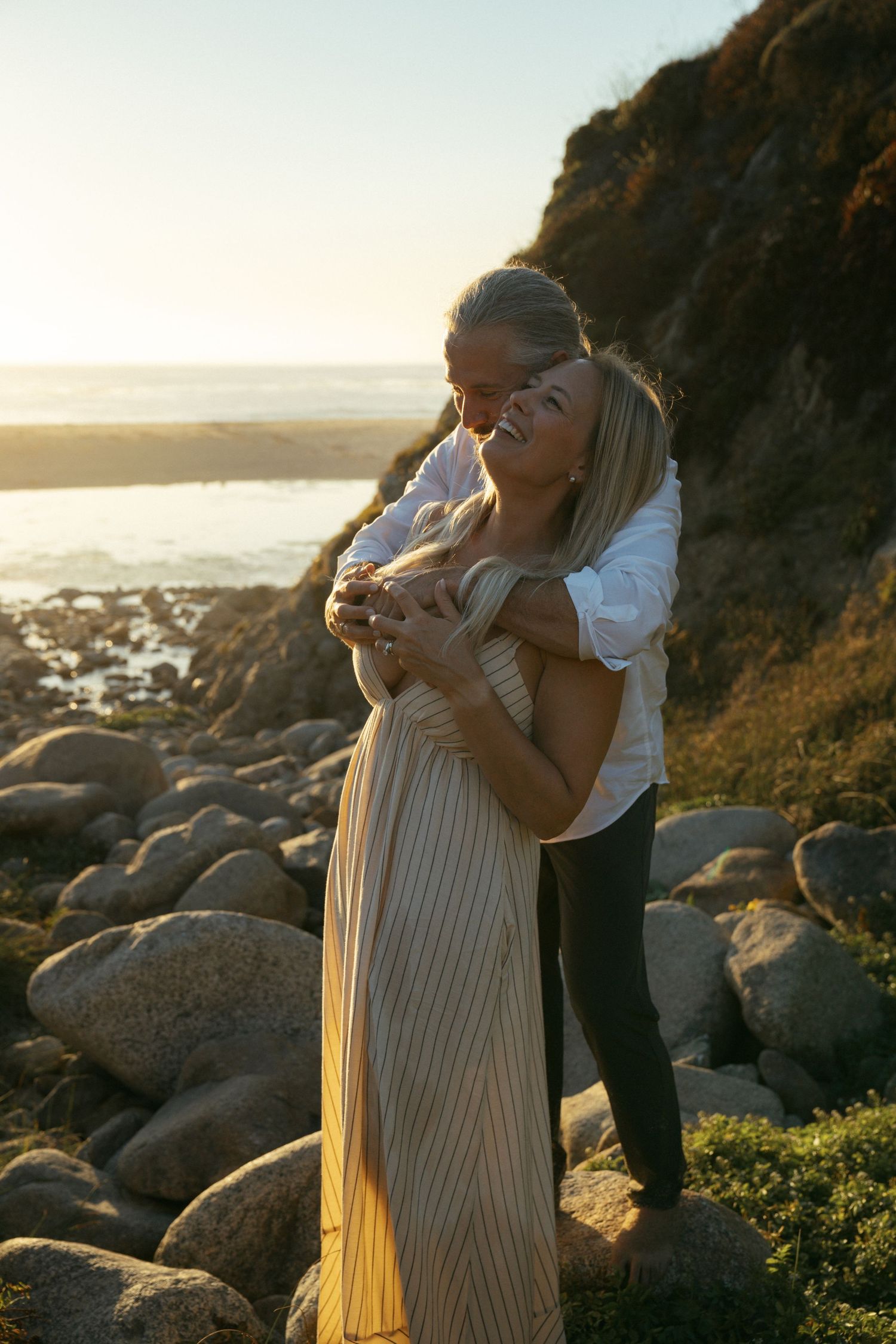 Couple sharing an intimate moment on a rocky beach at sunset with ocean cliffs in the background.