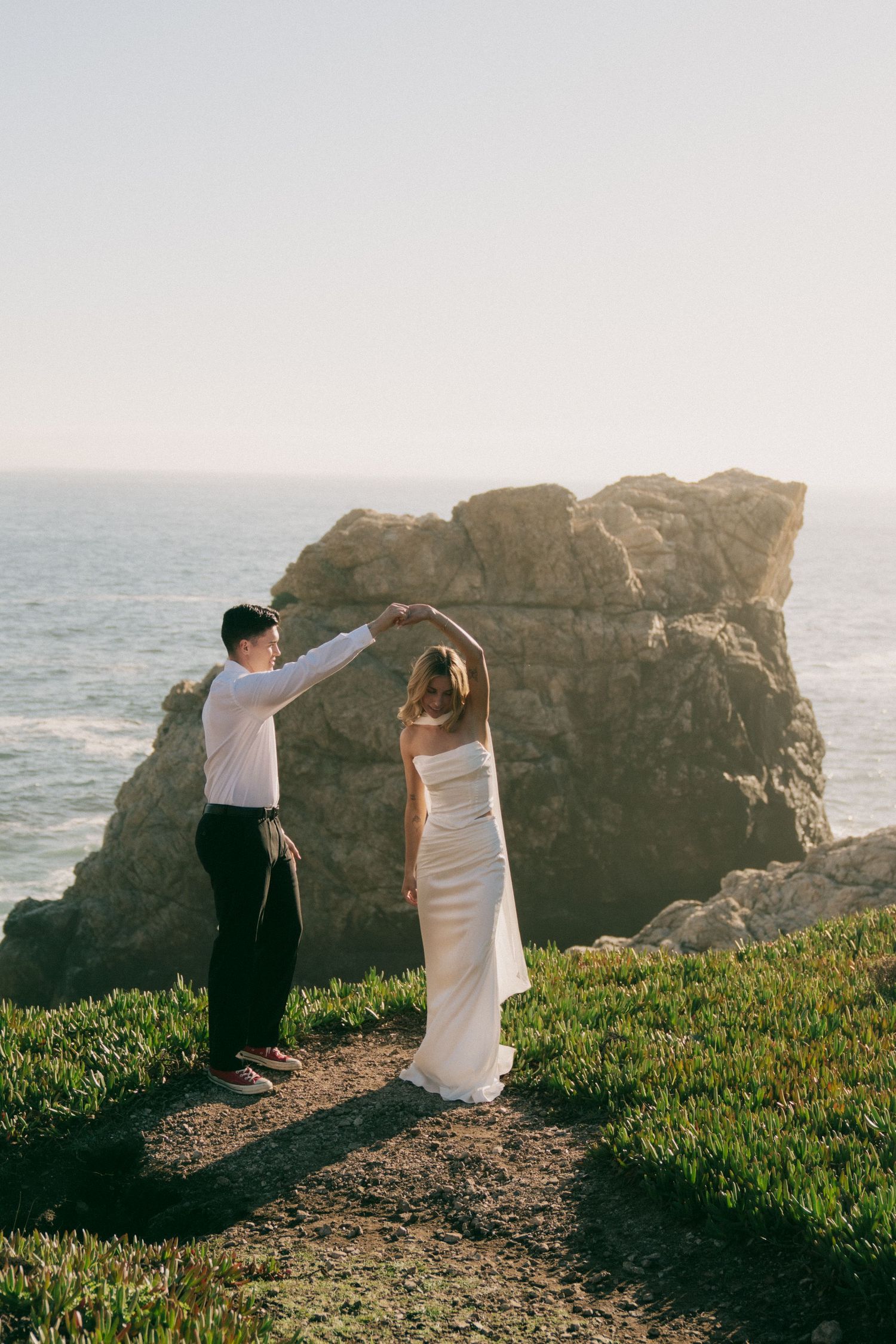 A couple dances together on a cliff overlooking the ocean during golden hour, with dramatic coastal rocks in the background.