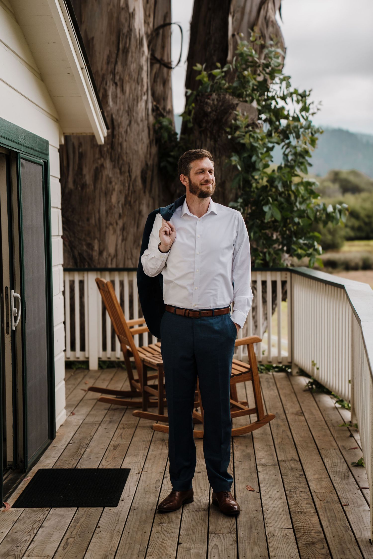 A person stands on a rustic wooden deck wearing business casual attire with rocking chairs and mountain views in background.