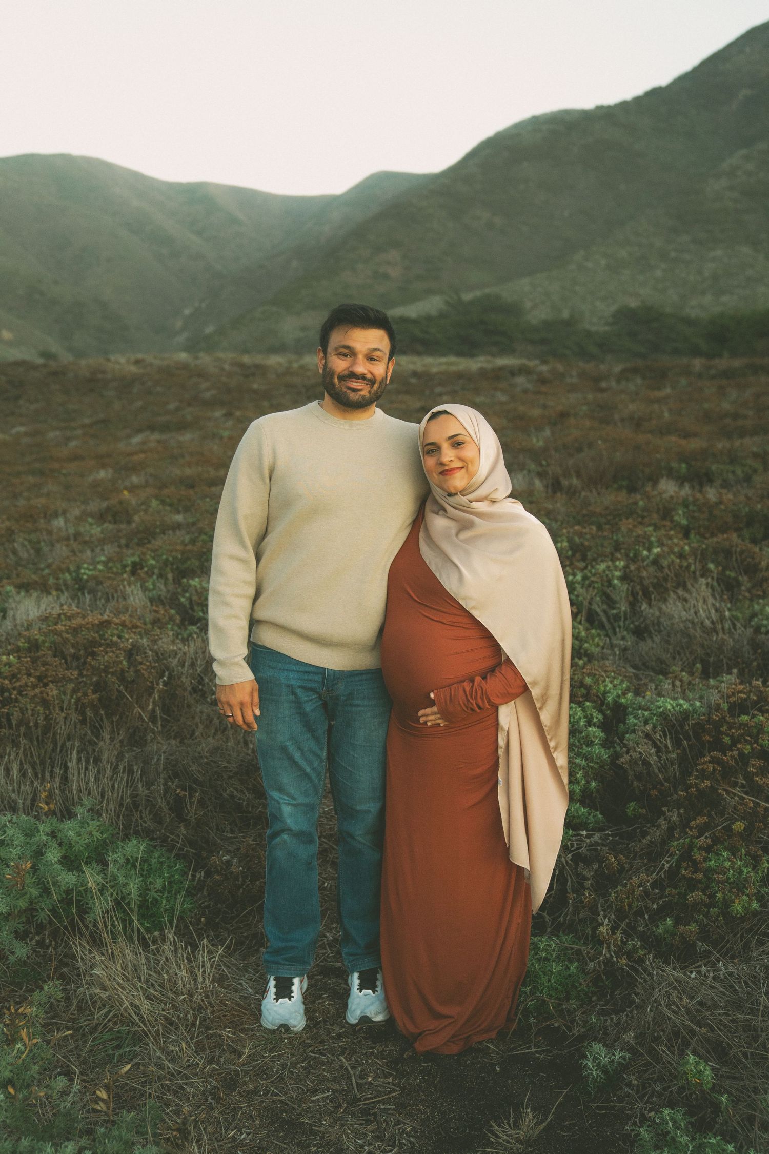 A couple stands together in a scenic mountainous landscape wearing casual clothing and warm smiles.