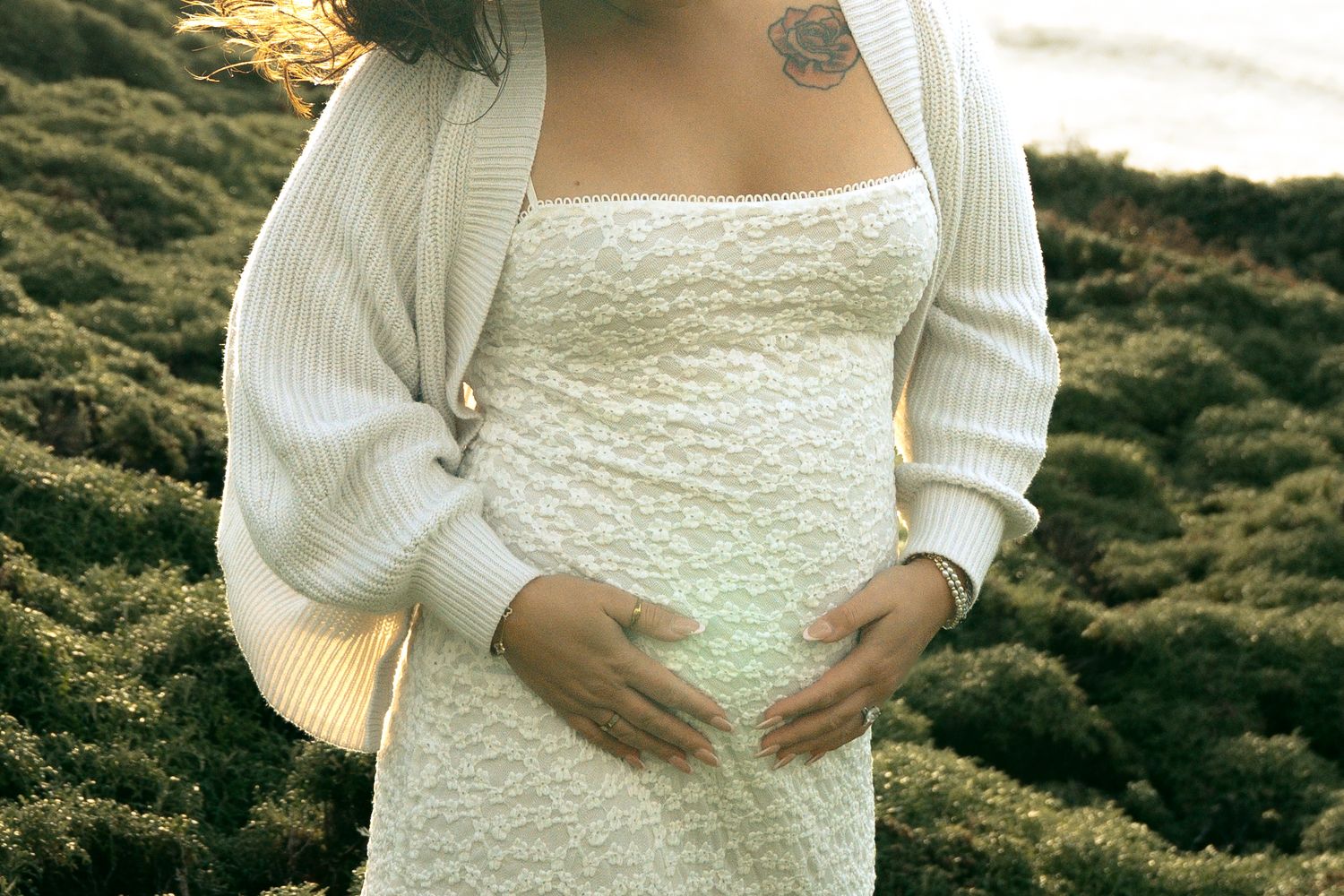 Pregnant woman in white lace dress and cream cardigan cradling baby bump outdoors on rocky terrain.
