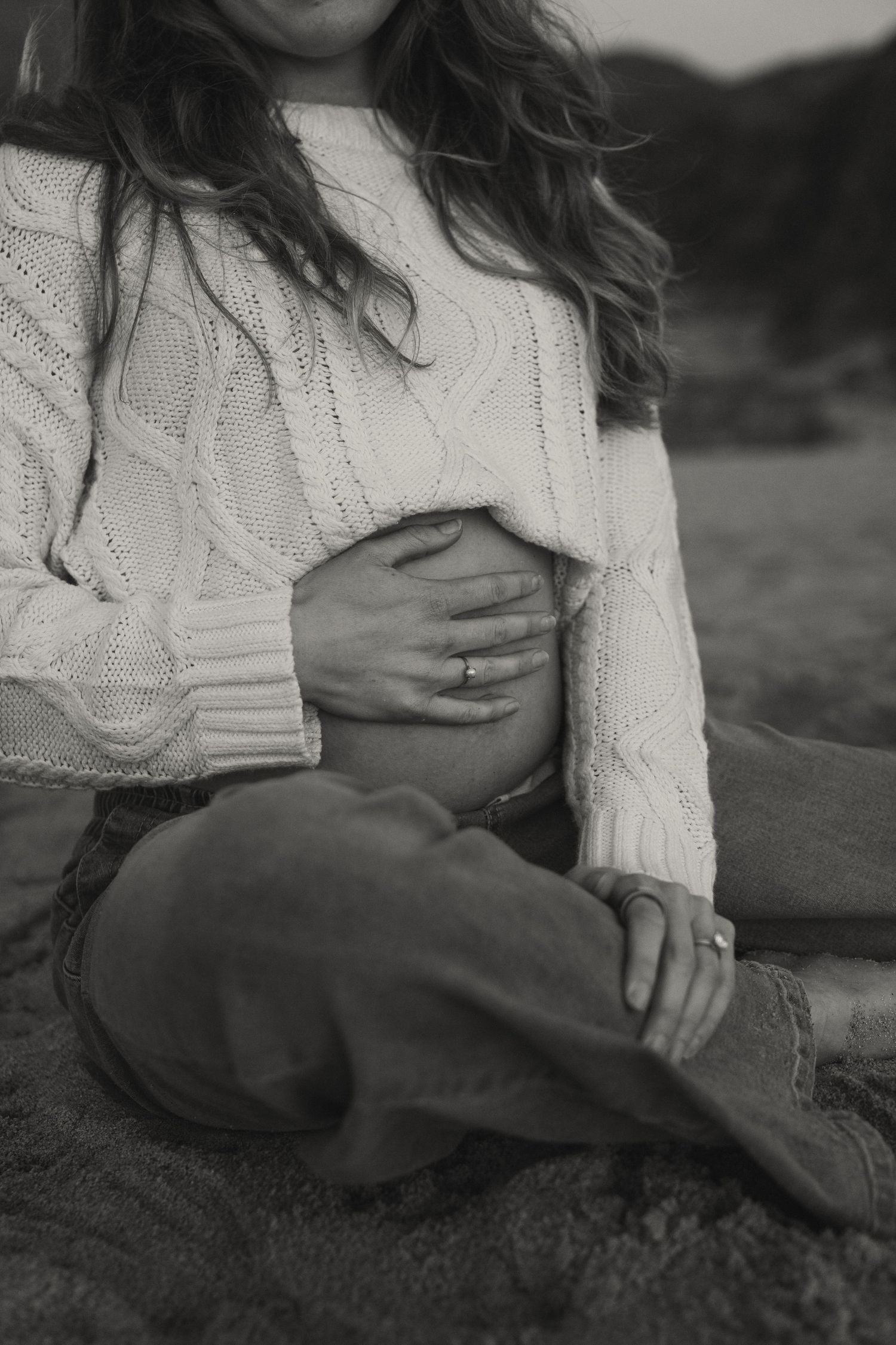 Pregnant woman in white cable knit sweater cradling baby bump while sitting on beach in black and white photo.