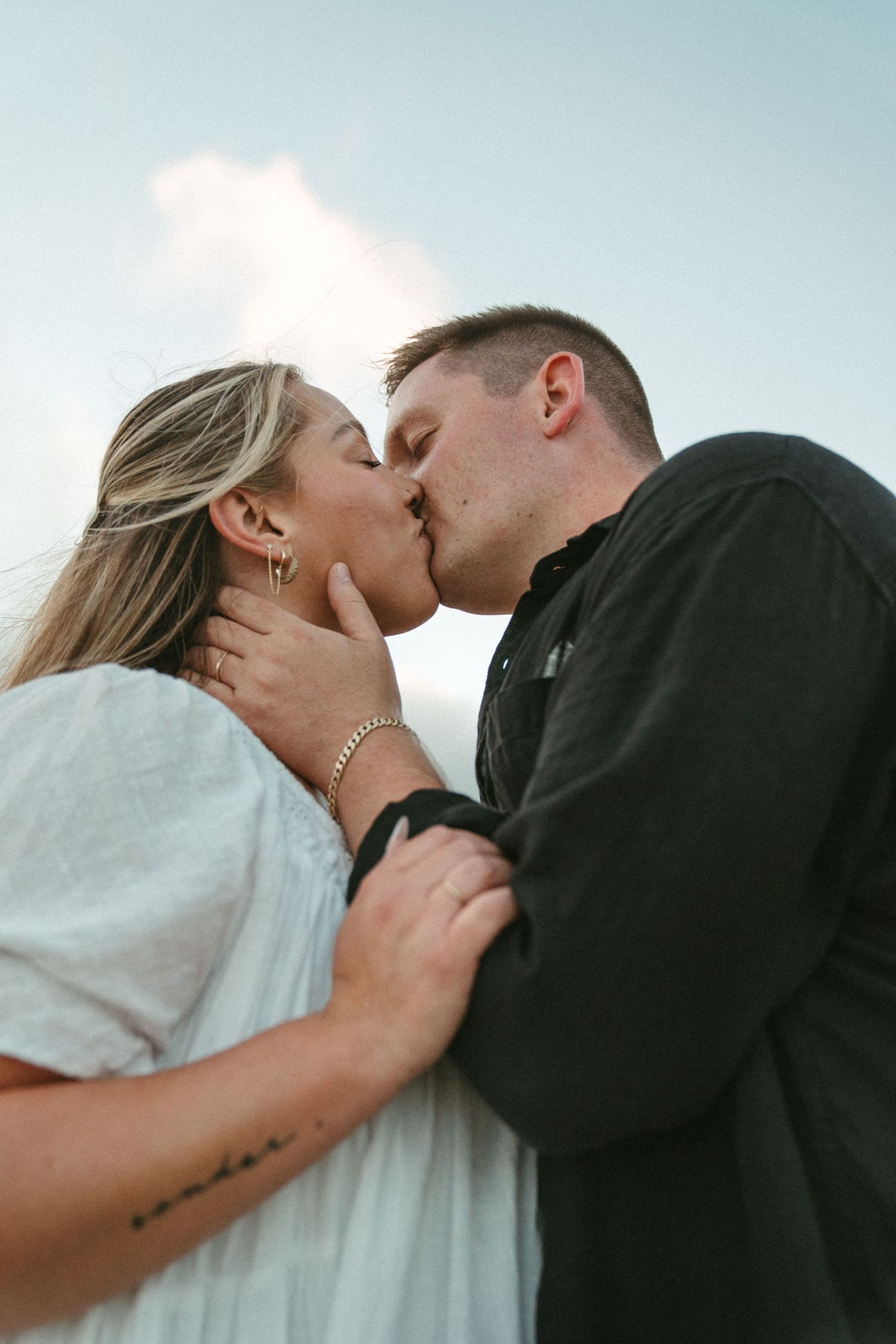 A couple shares a romantic kiss against a soft blue sky background.